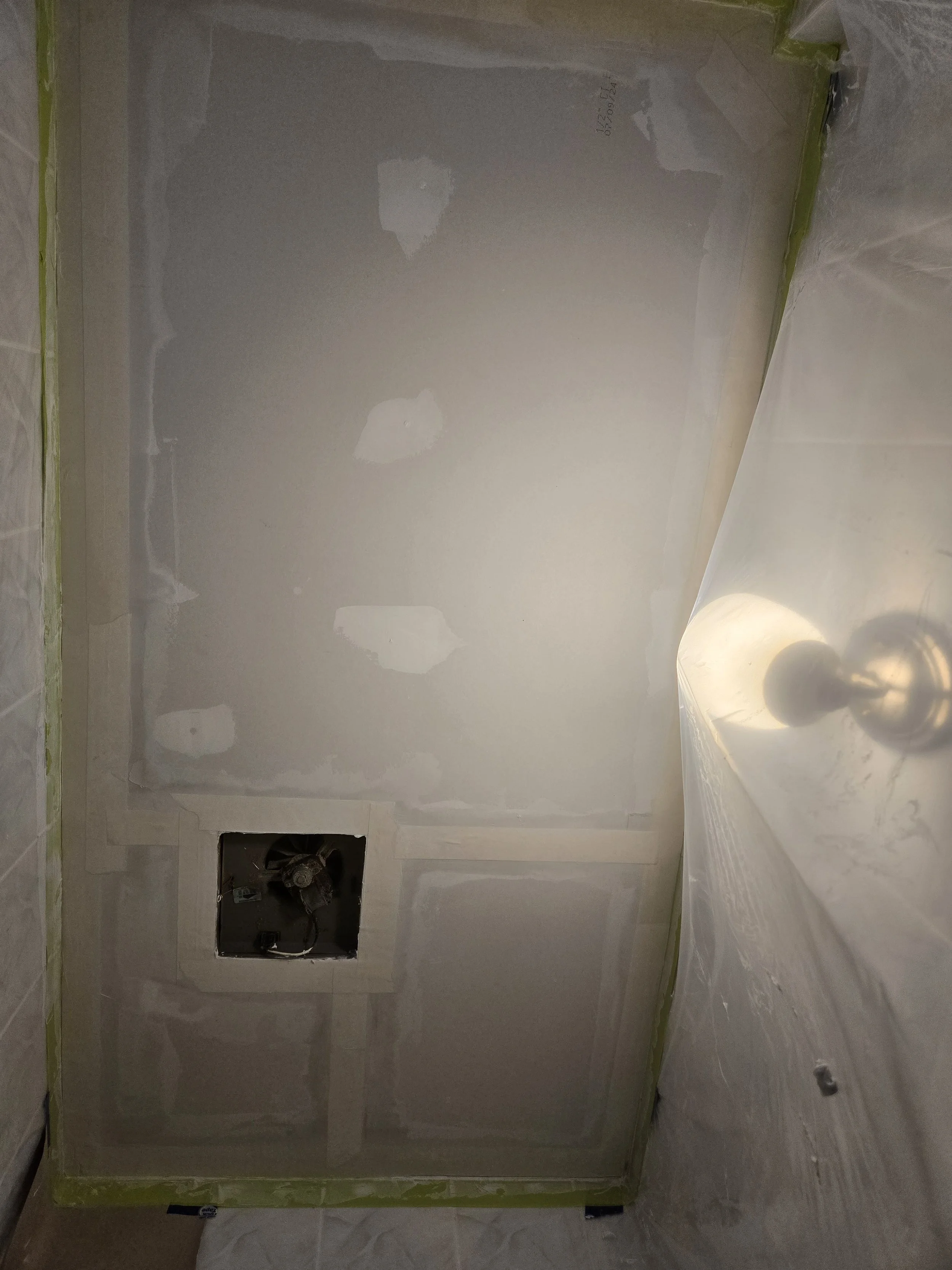 Bathroom under renovation showing drywall with patches, a vent duct opening, and a sink with a faucet wrapped in plastic, with painter's tape along the edges.