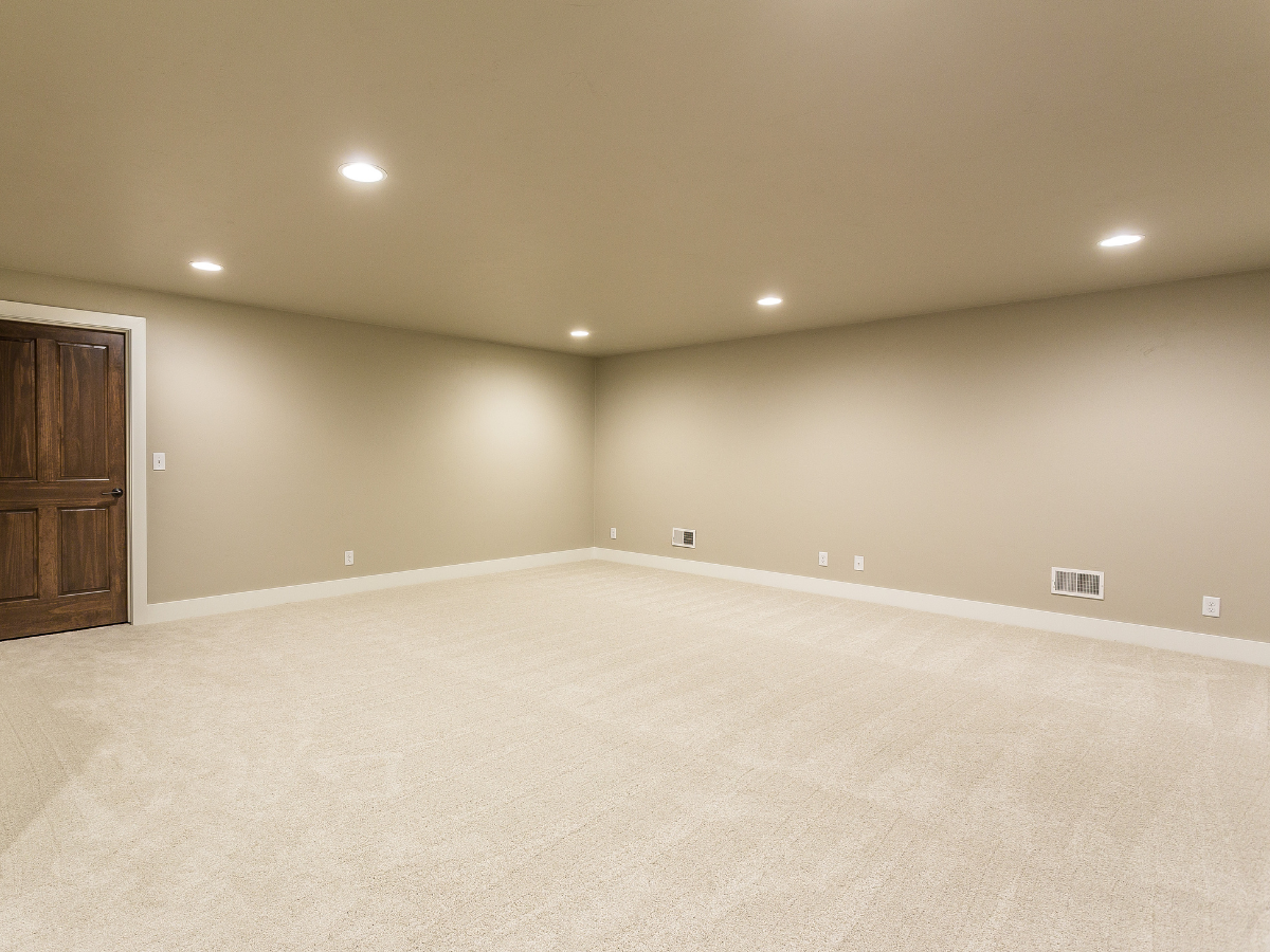 Empty beige room with a wooden door, beige carpet, and ceiling lights.