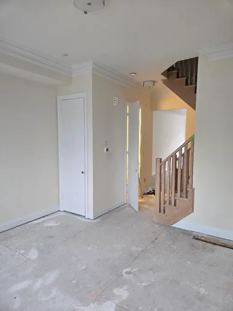 Empty room with white walls, a closed door, open door revealing a lit hallway, and wooden staircase.