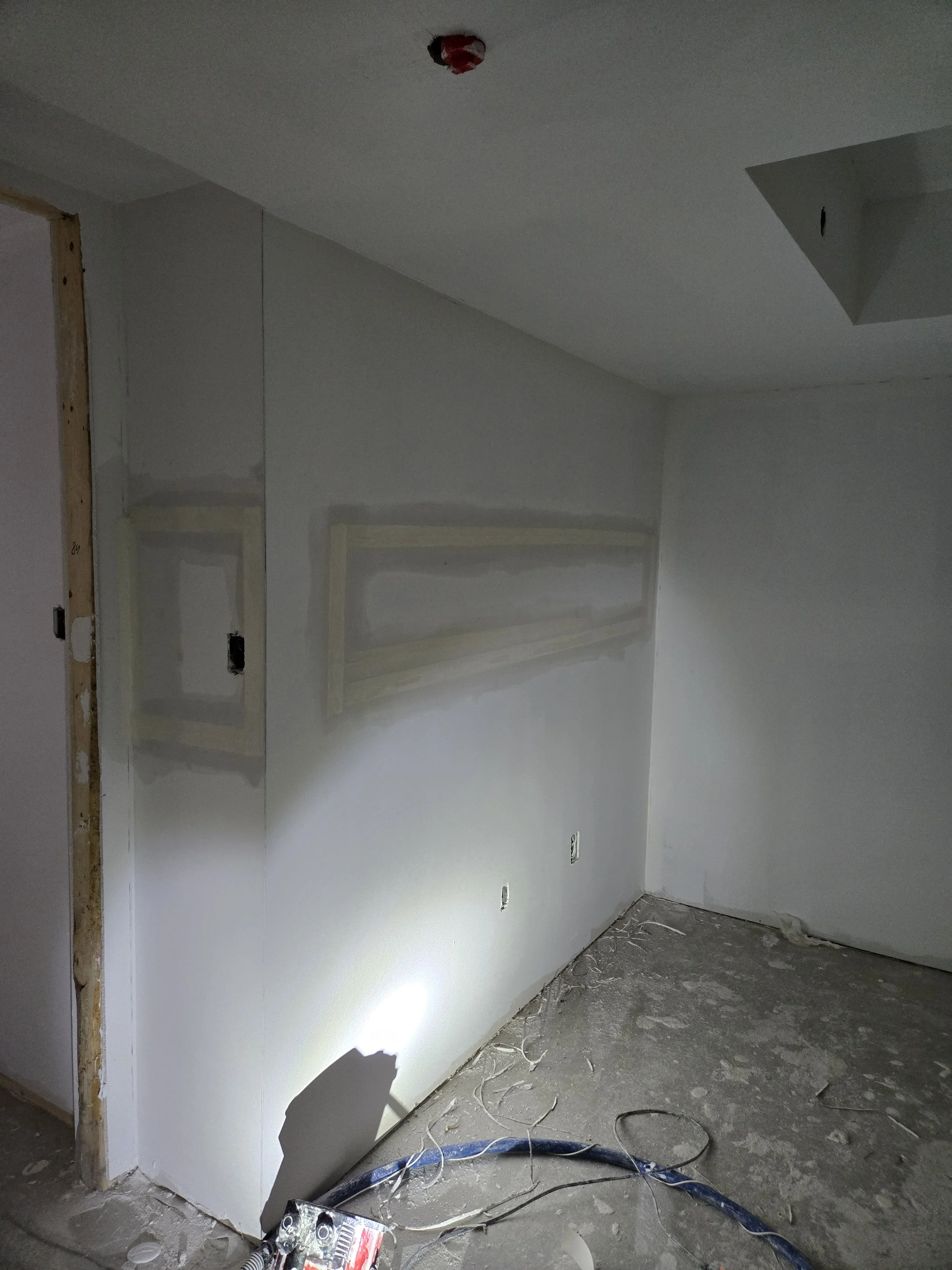 Interior of a room under construction with a white wall, electrical outlets, construction tools, and debris on the floor.