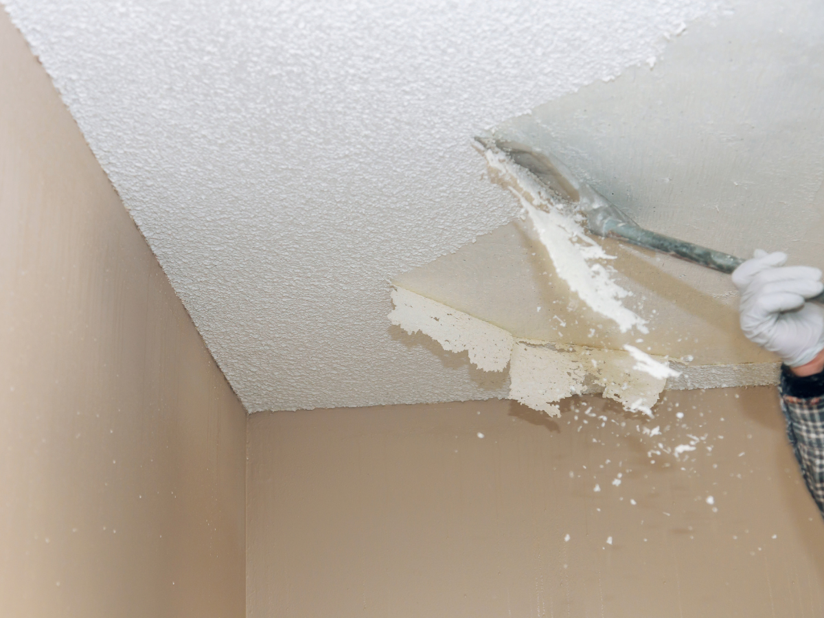 Person scraping textured ceiling with a putty knife, wearing white gloves and a plaid long sleeve shirt.