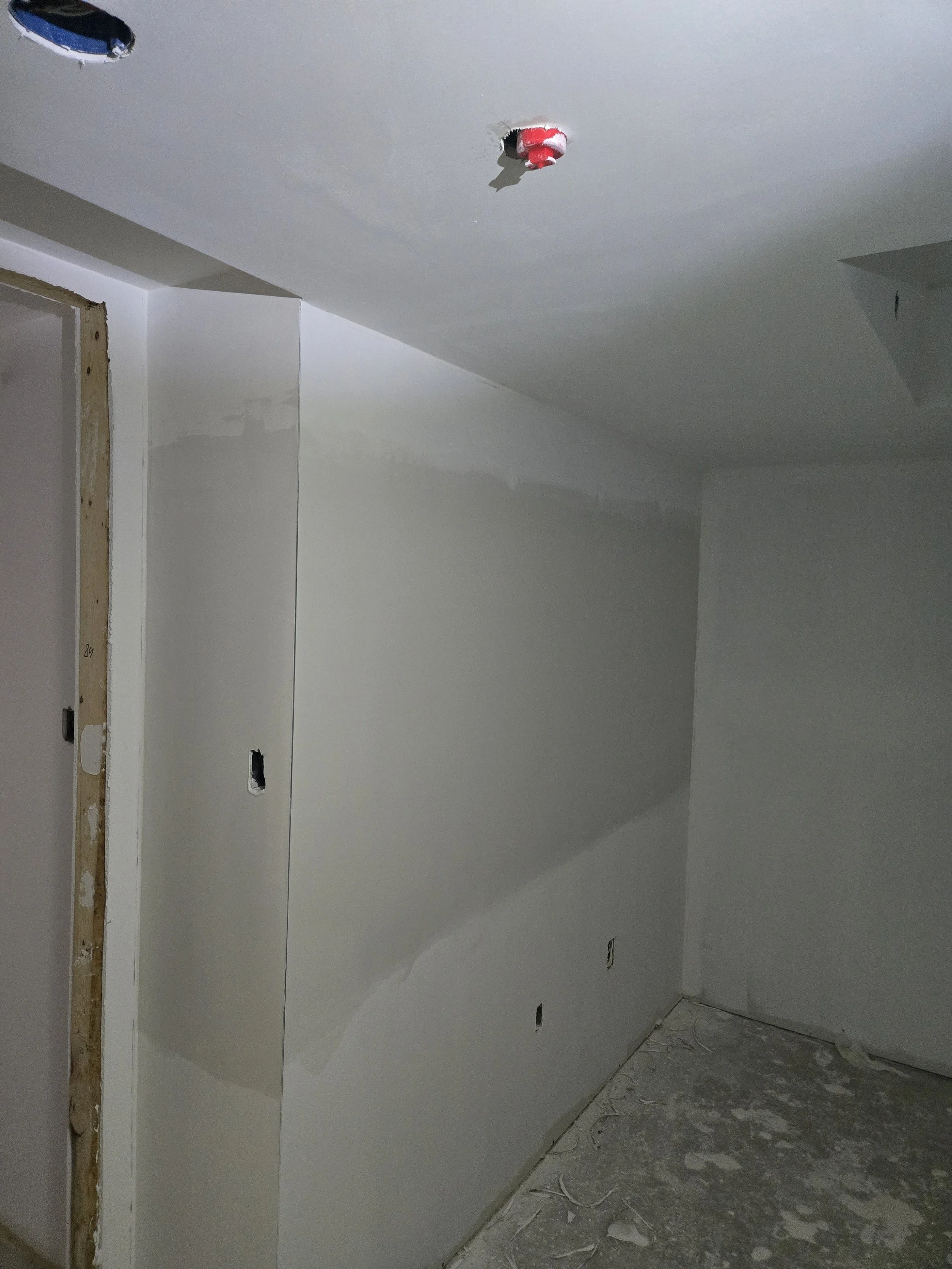 Interior view of a room under construction or renovation, with drywall partially installed on the walls, unfinished edges, exposed electrical outlets, and construction debris on the floor.