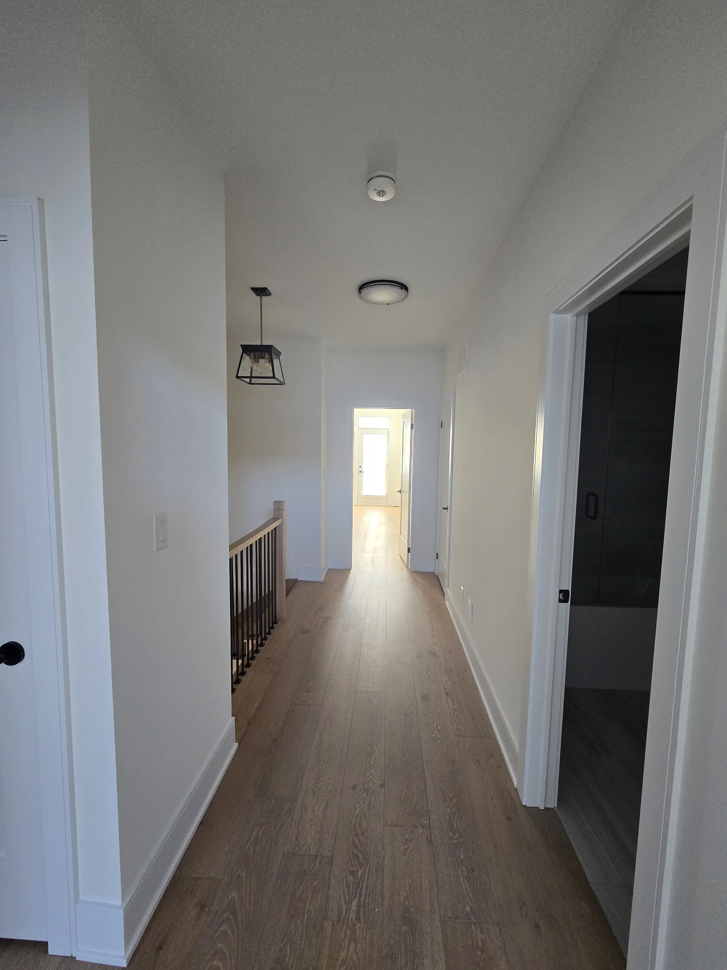 Interior of a hallway with white walls and wooden flooring. There are multiple doors and a staircase with a wooden handrail on the left. The ceiling has two light fixtures and a smoke detector. At the end of the hallway, there is a door with a window