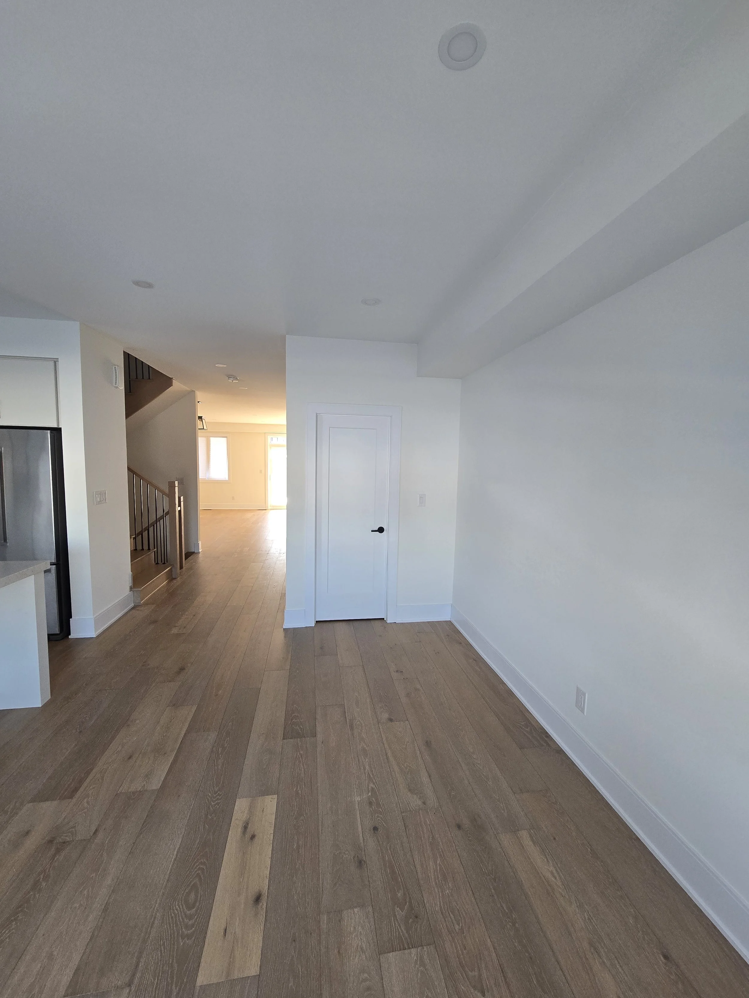 Interior of a house showing a room with wooden flooring, white walls, and a door. The background displays a staircase with a wooden handrail and open space filled with natural light.