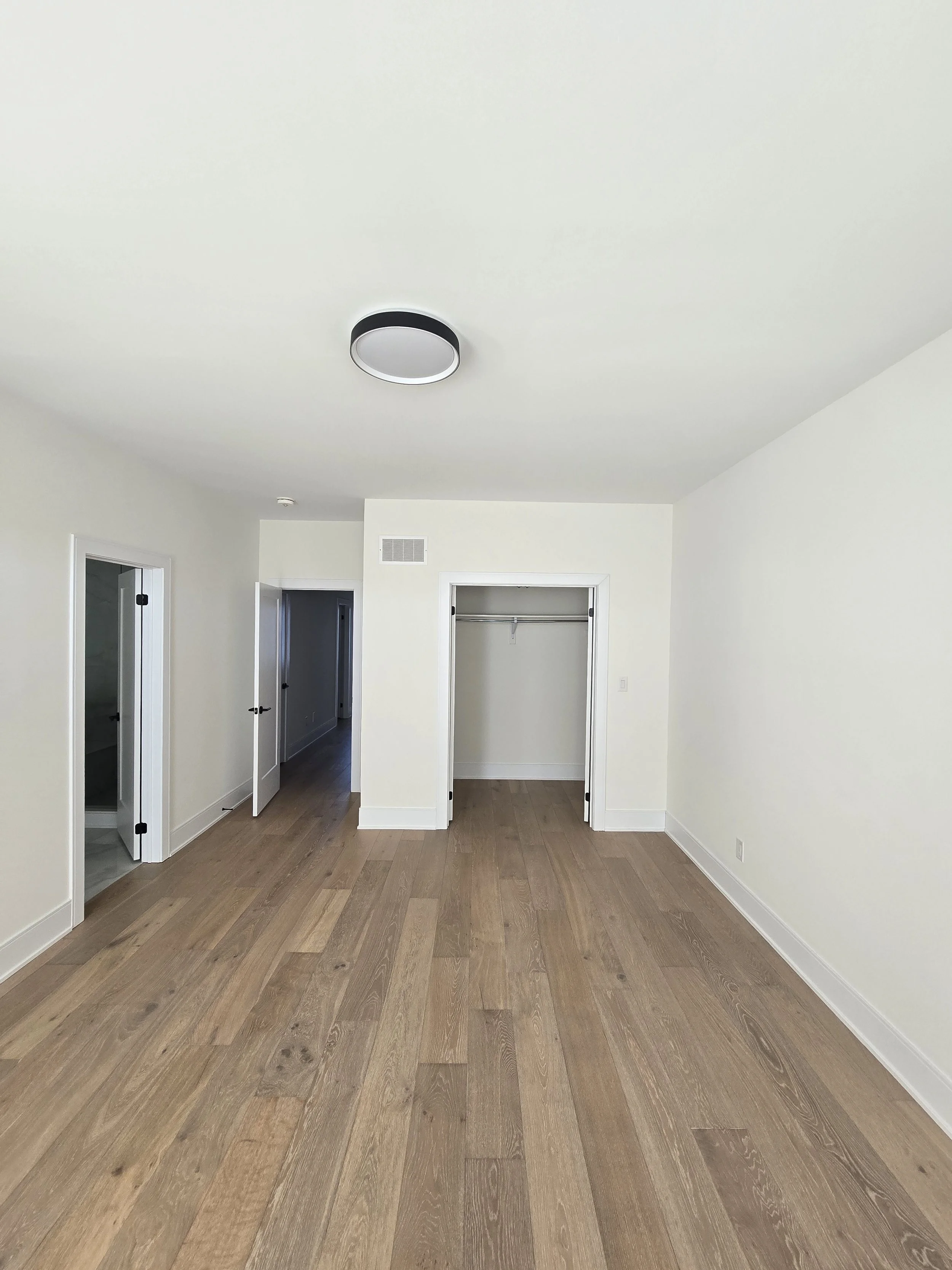 Empty room with beige walls, light wood flooring, an open closet, and a ceiling light fixture.