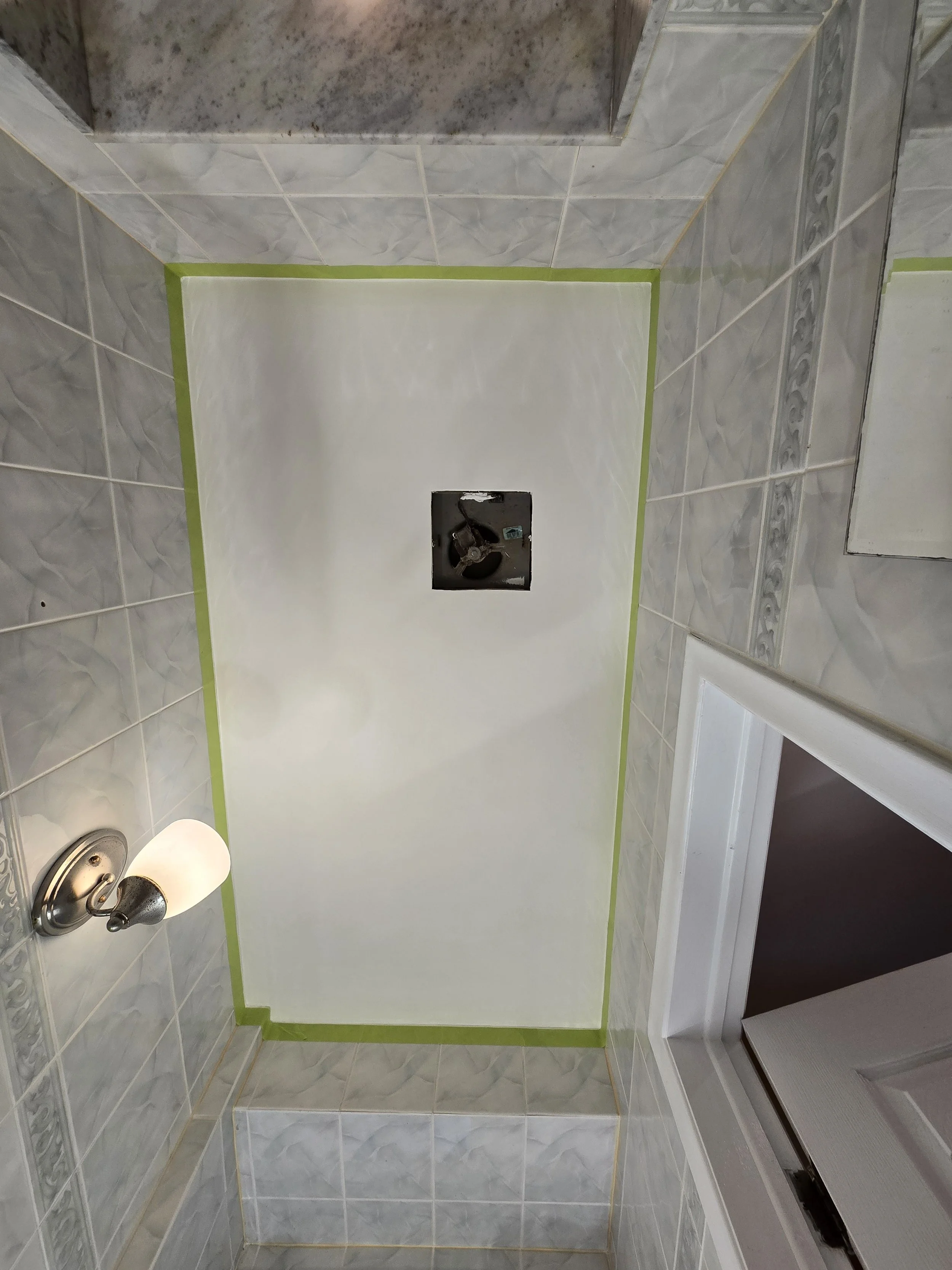 A partially tiled bathroom or shower area with a small, square, black opening in the wall for plumbing. The surrounding walls are tiled with white and gray marble-patterned tiles. Green painter's tape outlines a section on the wall, possibly for ongo