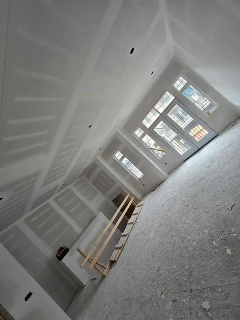 Interior of an unfinished room with drywall, windows, and construction materials, including a small wooden workbench and scaffolding.