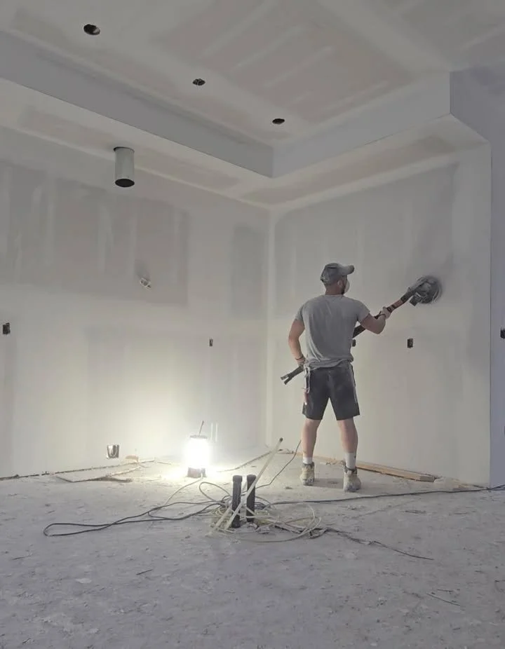 A worker using a power sander to smooth drywall on a wall during construction, with construction tools and wires on the unfinished floor and ceiling lights not yet installed.