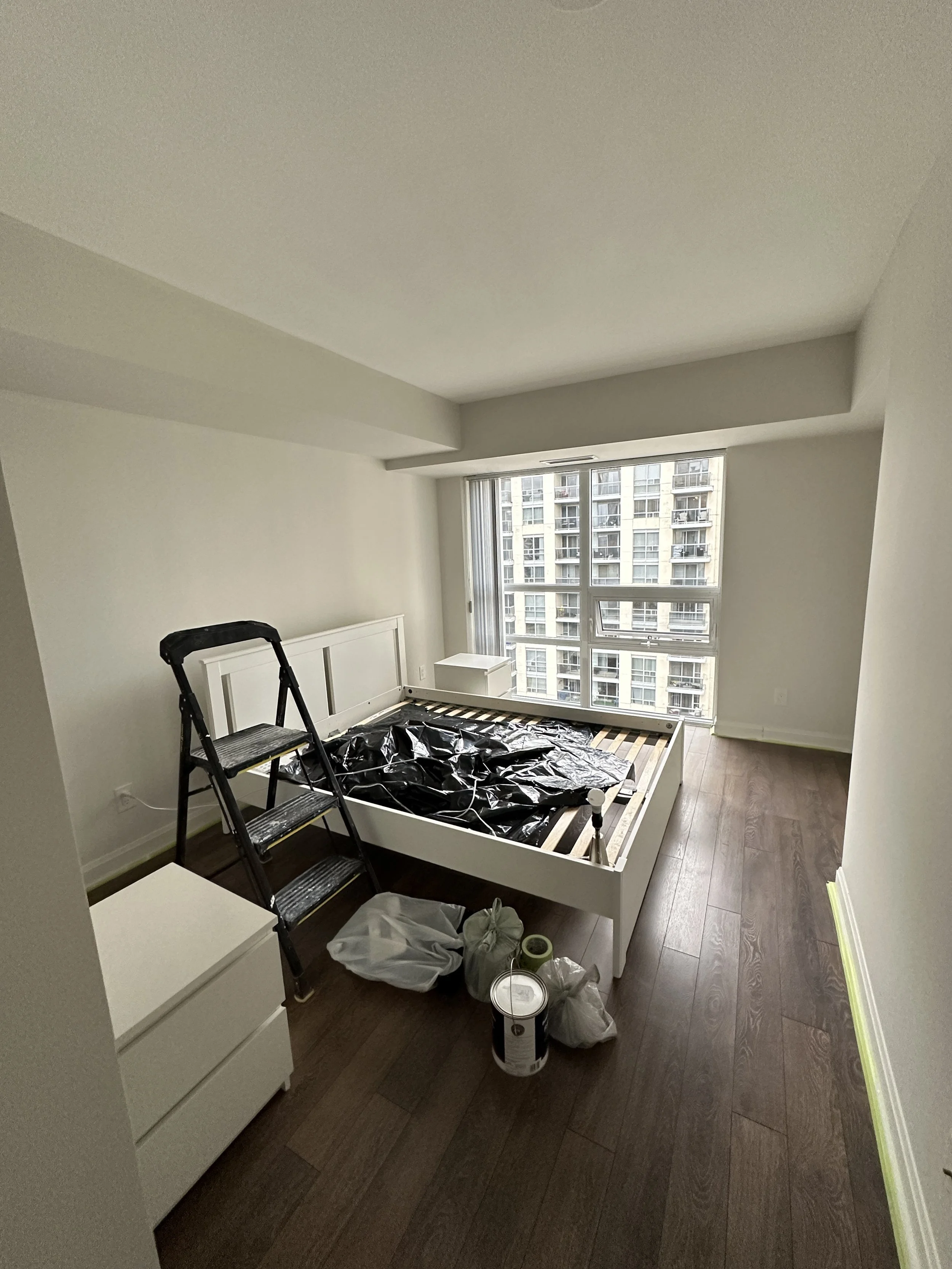 Unfinished bedroom with a bed frame, ladder, paint supplies, window view of a tall apartment building, and some scattered household items.