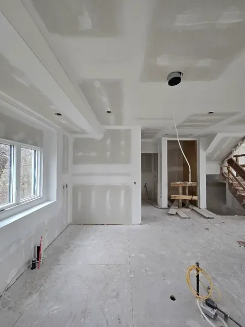Interior of a room under construction with drywall installed, a window on the left, and construction equipment and wiring visible.