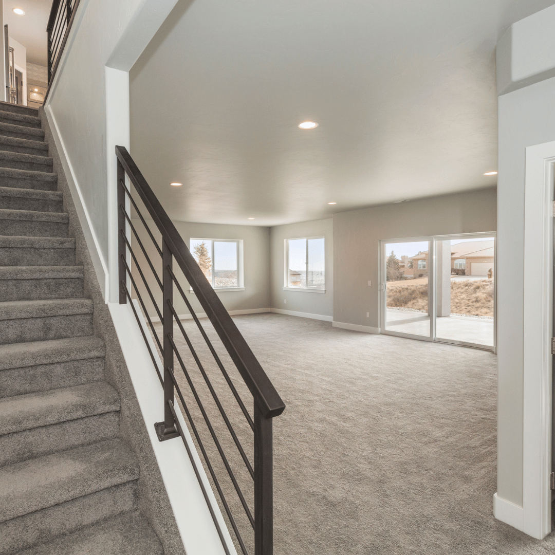 Empty living room with carpeted floor, multiple large windows, sliding glass door leading to outside, and a staircase with dark railing on the left.