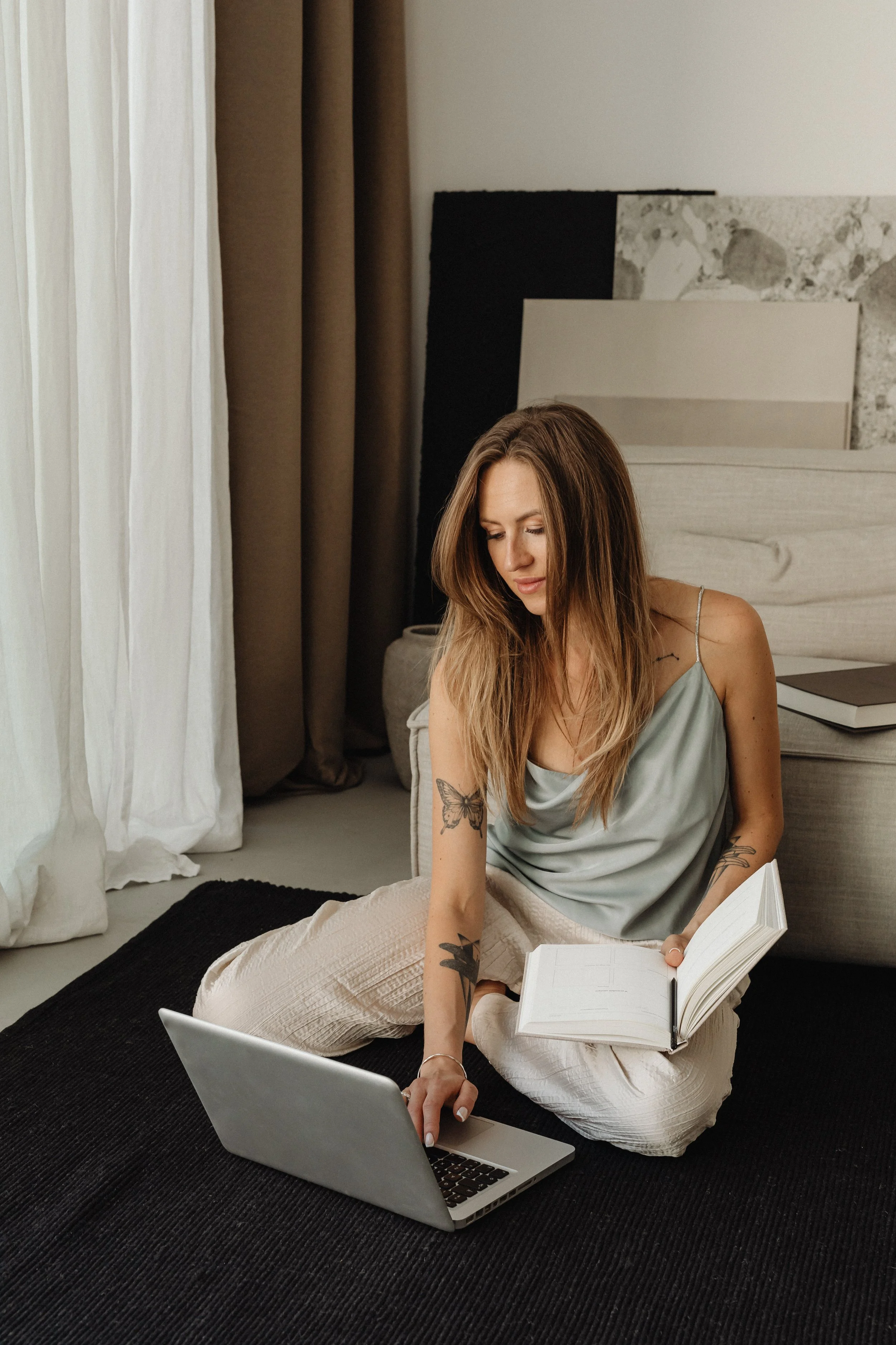 A woman with long hair and tattoos on her arm, wearing a silver top and beige pants, sitting cross-legged on a black rug, looking at a laptop, holding an open notebook, in a modern bedroom with curtains, a bed, and decorative panels in the background.