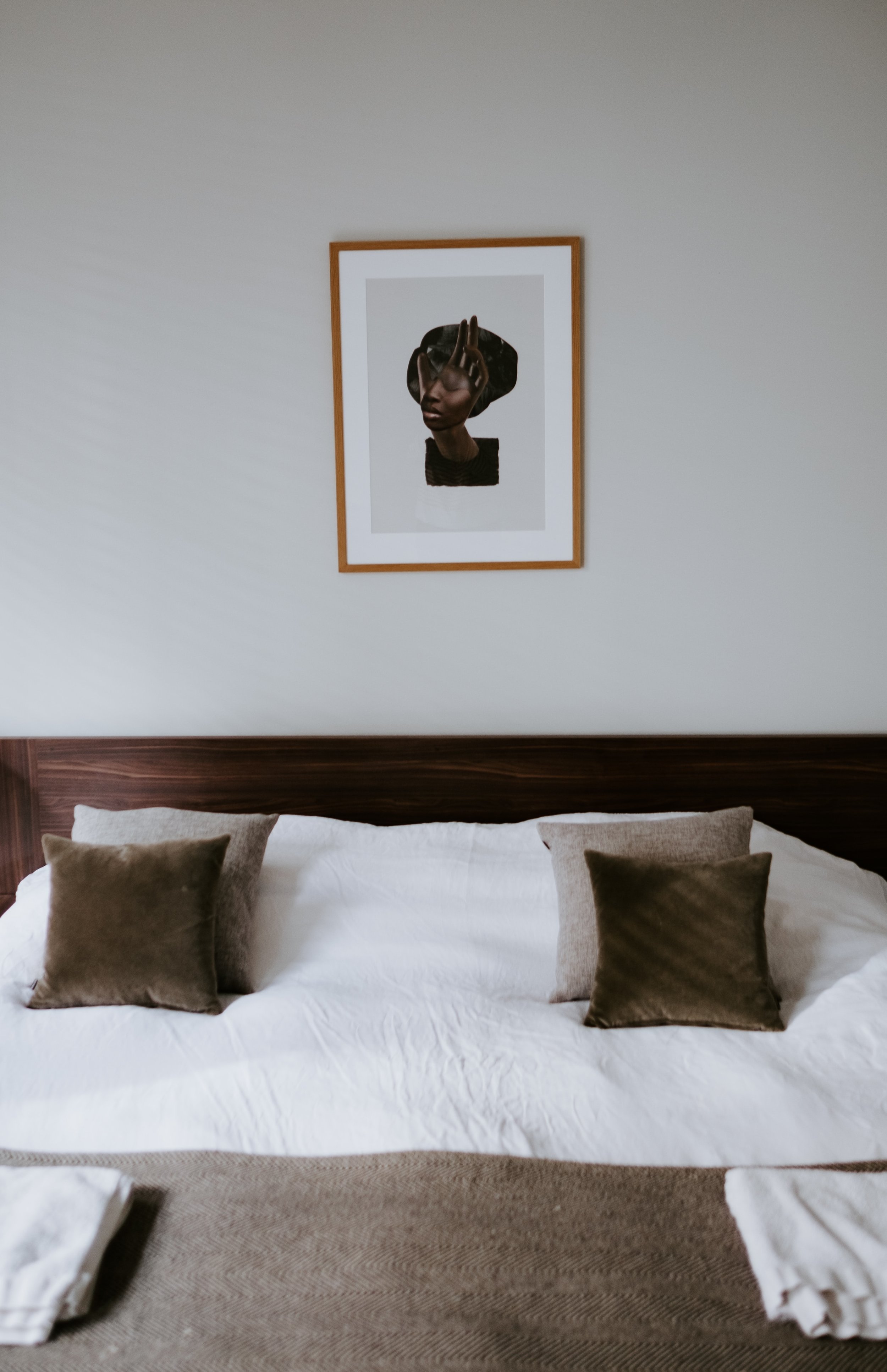 A neatly made bed with white sheets, beige and brown pillows, a wooden headboard, and a framed artwork of a woman on the white wall above.
