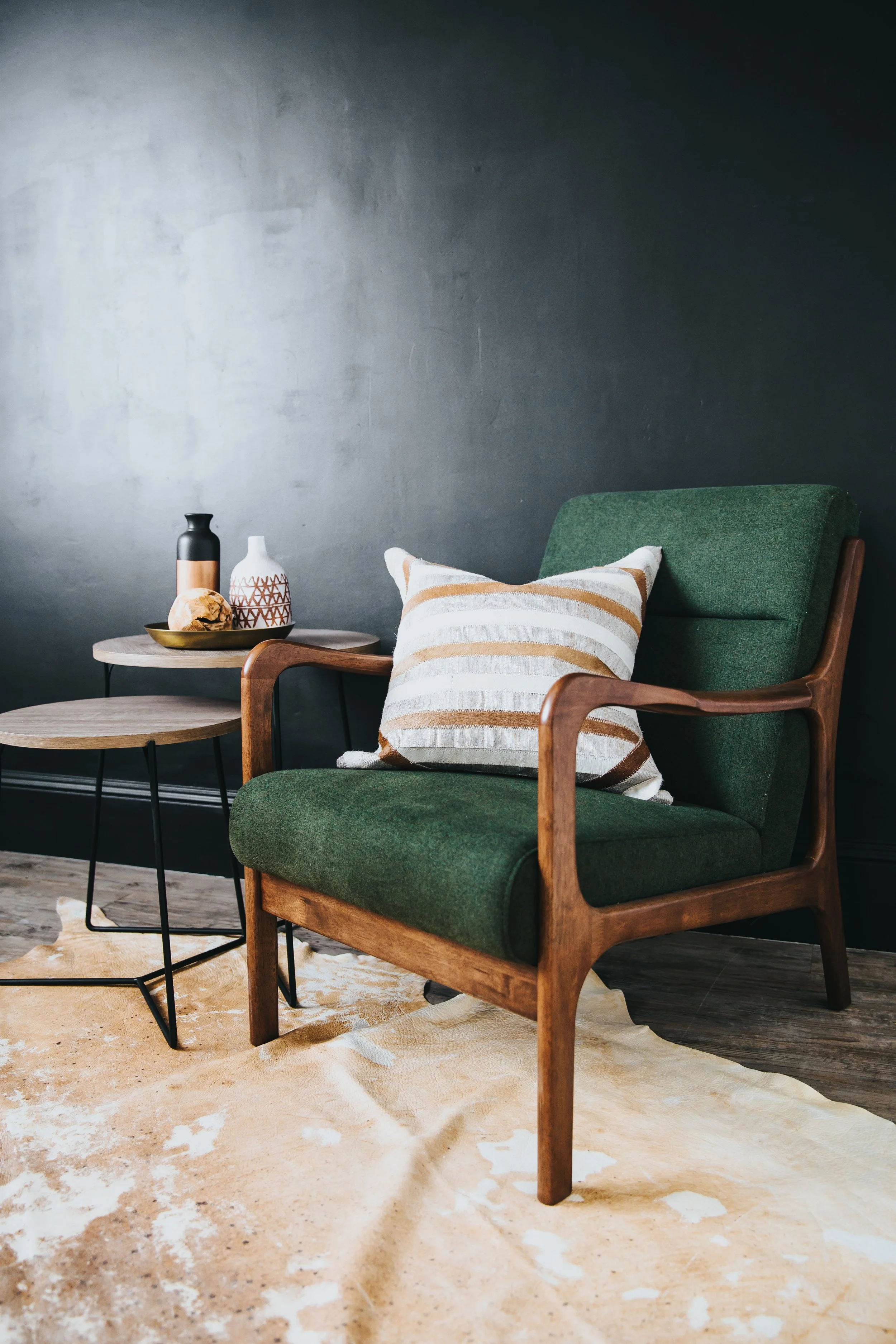 A green armchair with wooden frame, a beige and white striped pillow, a small wooden side table with black metal legs, decorative vases, a gold tray, and a cowhide rug in a room with dark gray wall.