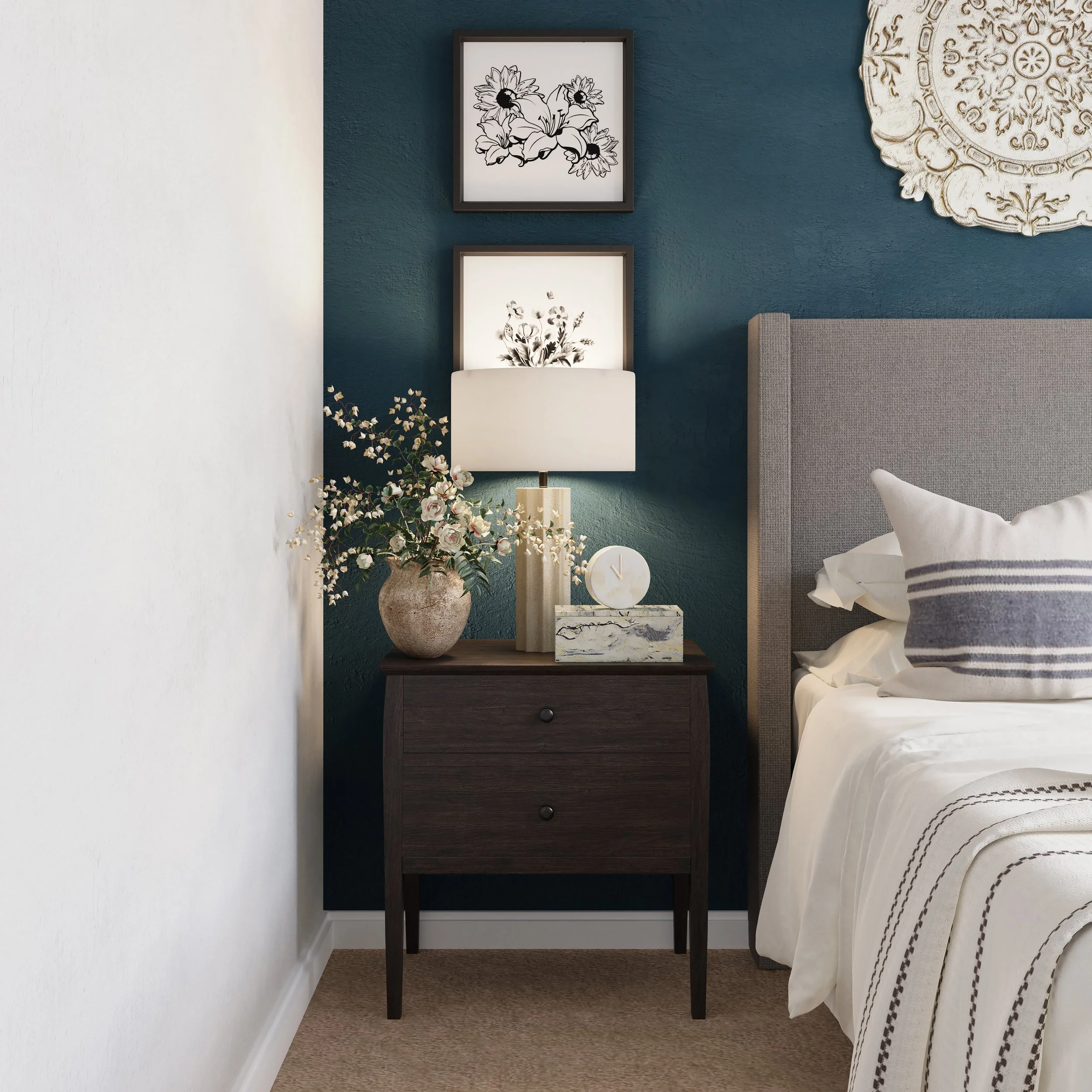 A bedroom corner with a black wooden nightstand, a white table lamp, a tall vase with pink and white flowers, and framed artwork of flowers on a dark teal wall.