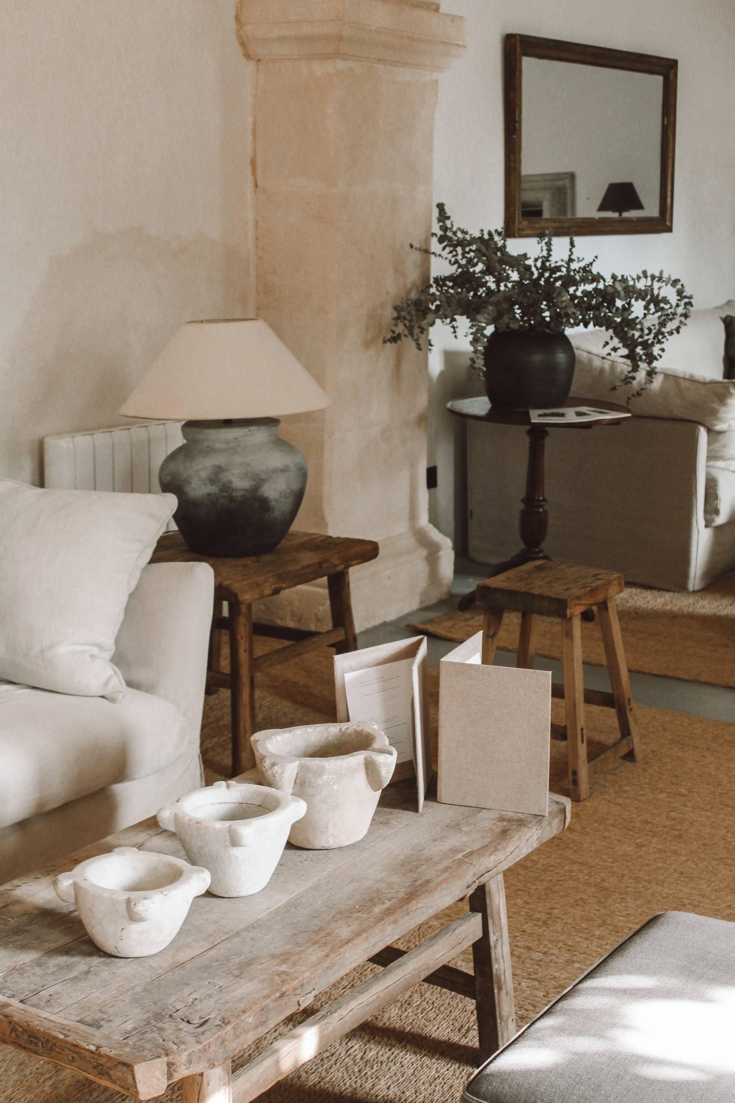A rustic wooden table displaying several white ceramic bowls. The scene is part of a cozy interior with a beige couch, a large black vase with greenery, a mirror on the wall, and some decorative boxes or books. The color palette includes neutral tone