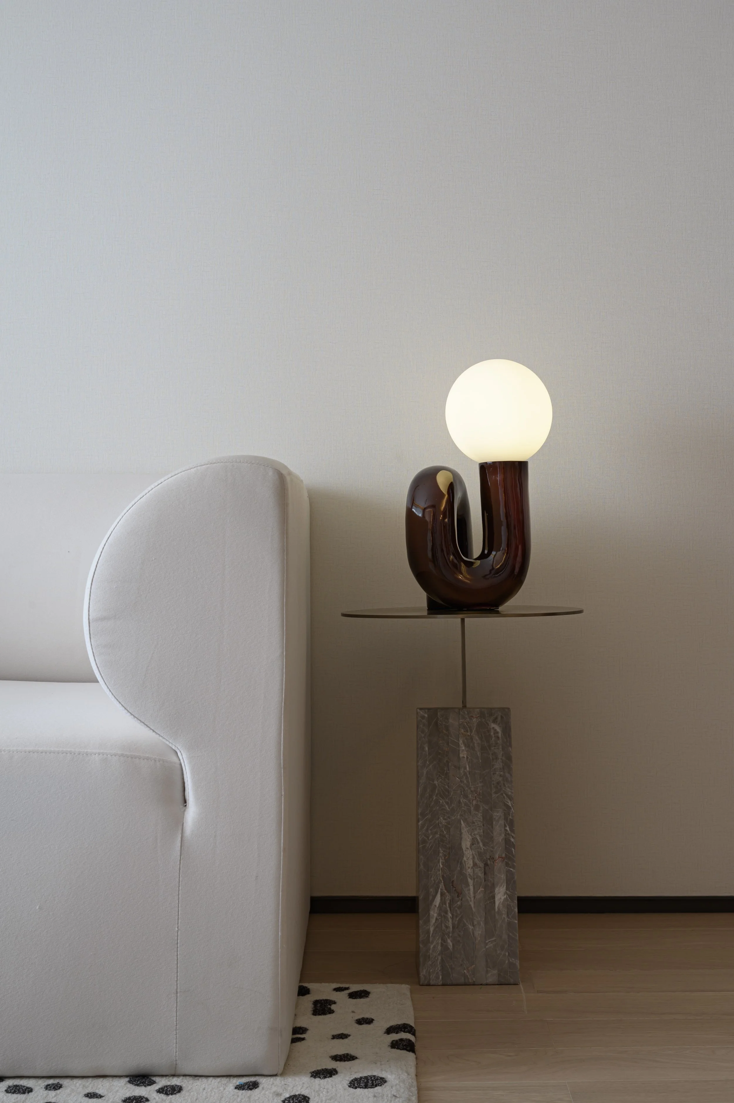 Photo of a white sofa, a bedside table with a marble base, and a decorative lamp with a ceramic base and a glowing white spherical shade, in a minimalist interior.