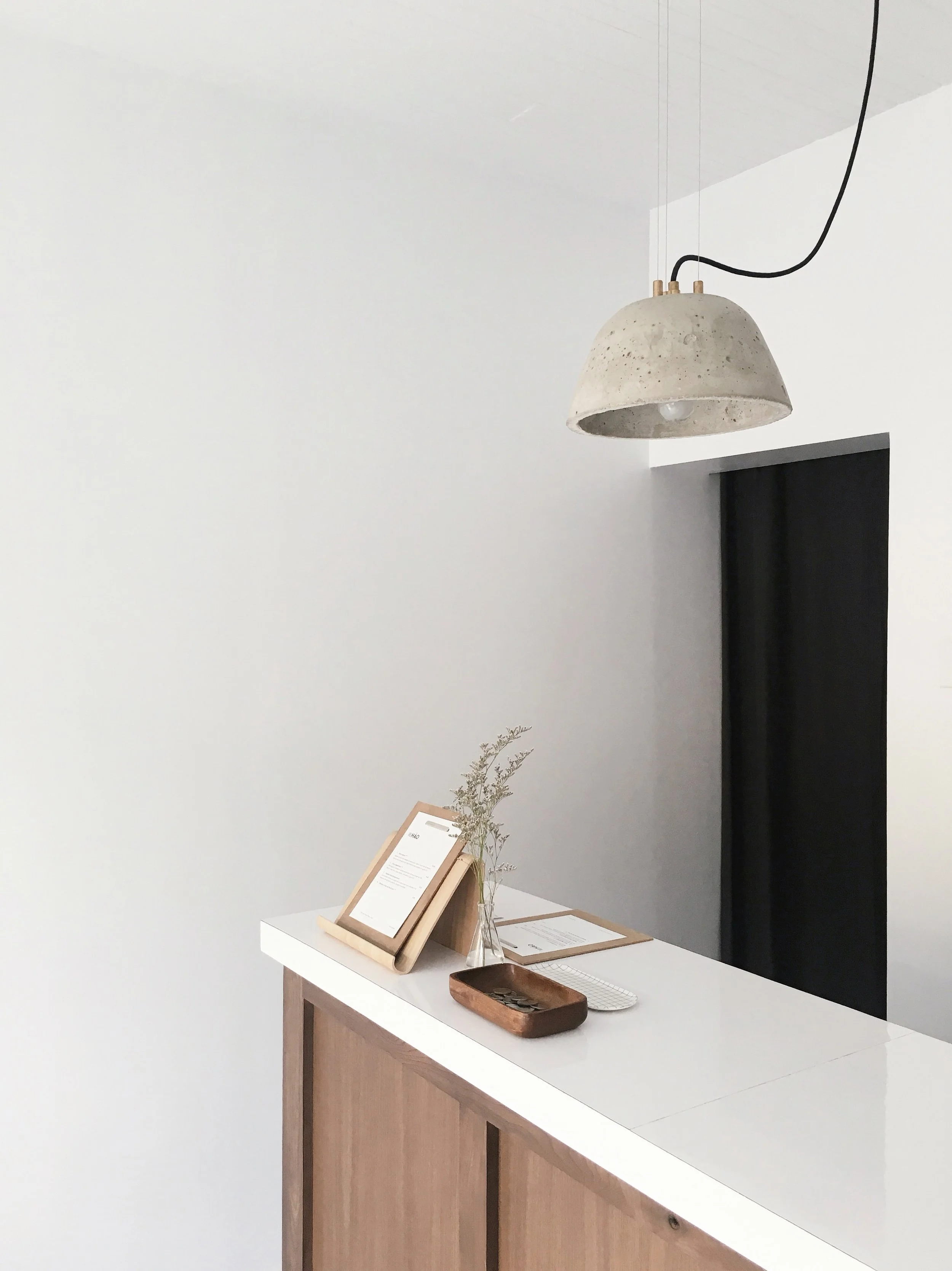 Minimalist interior scene with a wooden and white cabinet, a desk setup with a framed document, a glass vase with dried flowers, a wooden tray with coins, and a black window or door. A concrete pendant lamp hangs from the ceiling.