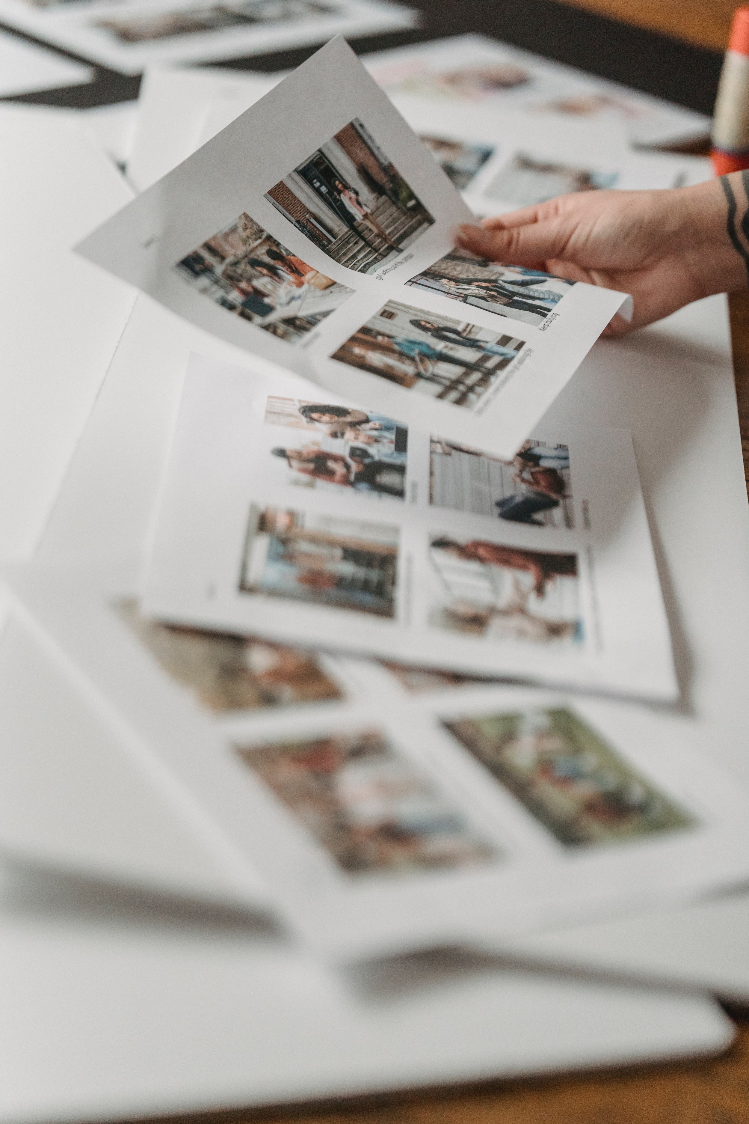 A person holding a sheet of photographs with a collection of small photos displayed on a white surface, some photos scattered around.