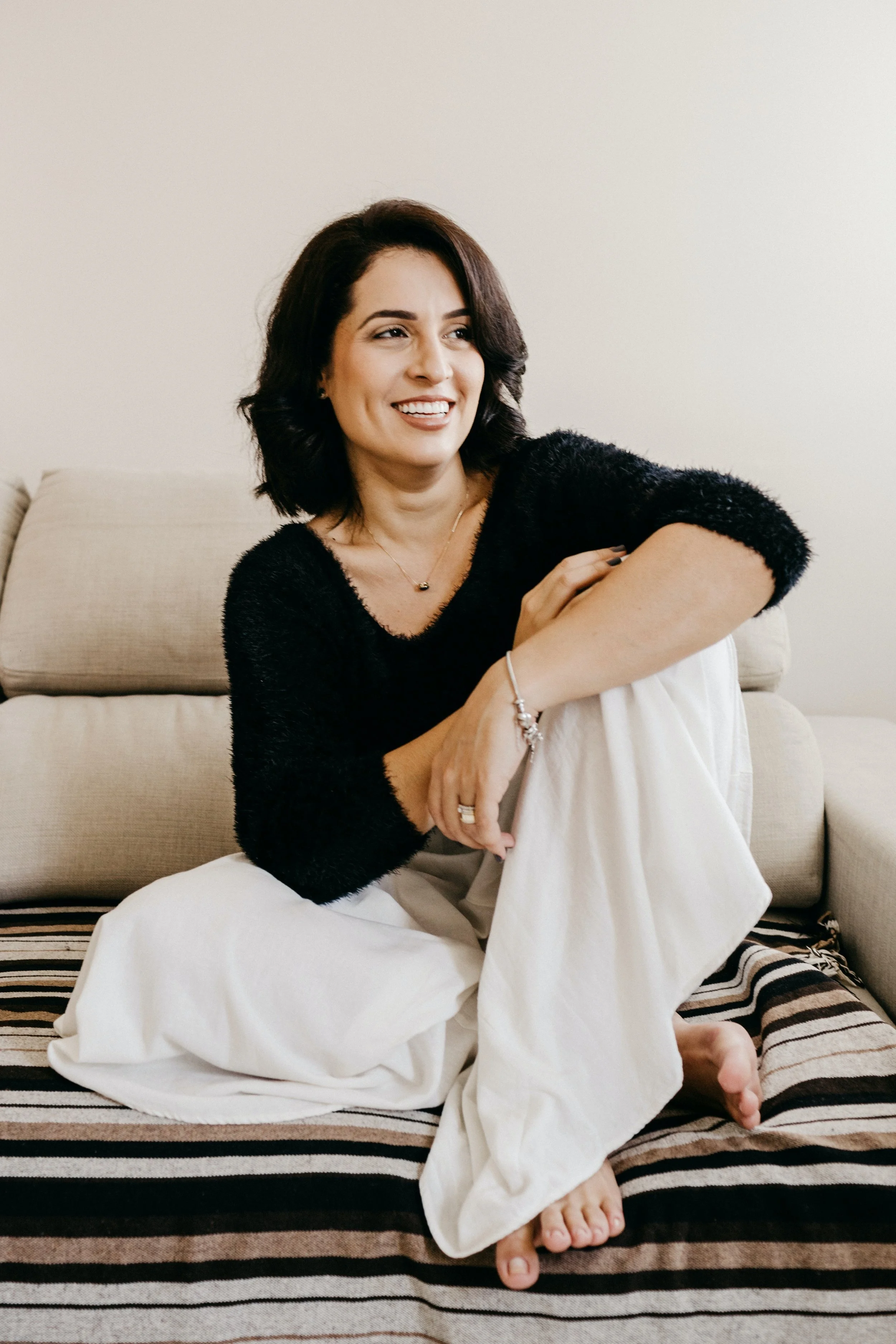 A woman with dark hair, wearing a black fuzzy sweater and white pants, sitting on a striped blanket on the floor, smiling, with her arms crossed and one leg folded, near a beige sofa in a room with plain light-colored walls.