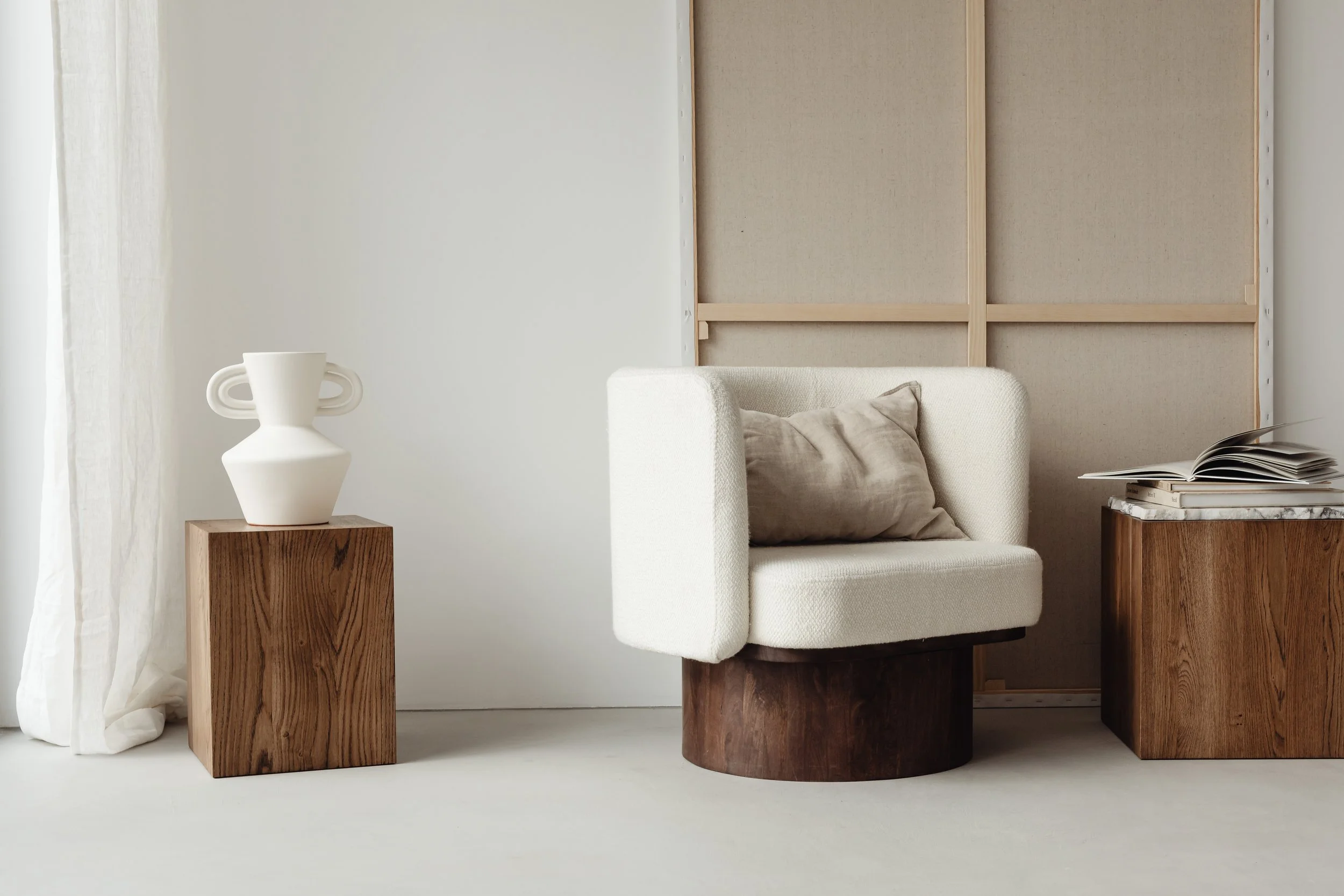Interior of a minimalist living space with a white sofa, beige wall panels with wooden frames, a small wooden side table with a white vase, a stack of books, and a beige throw pillow.