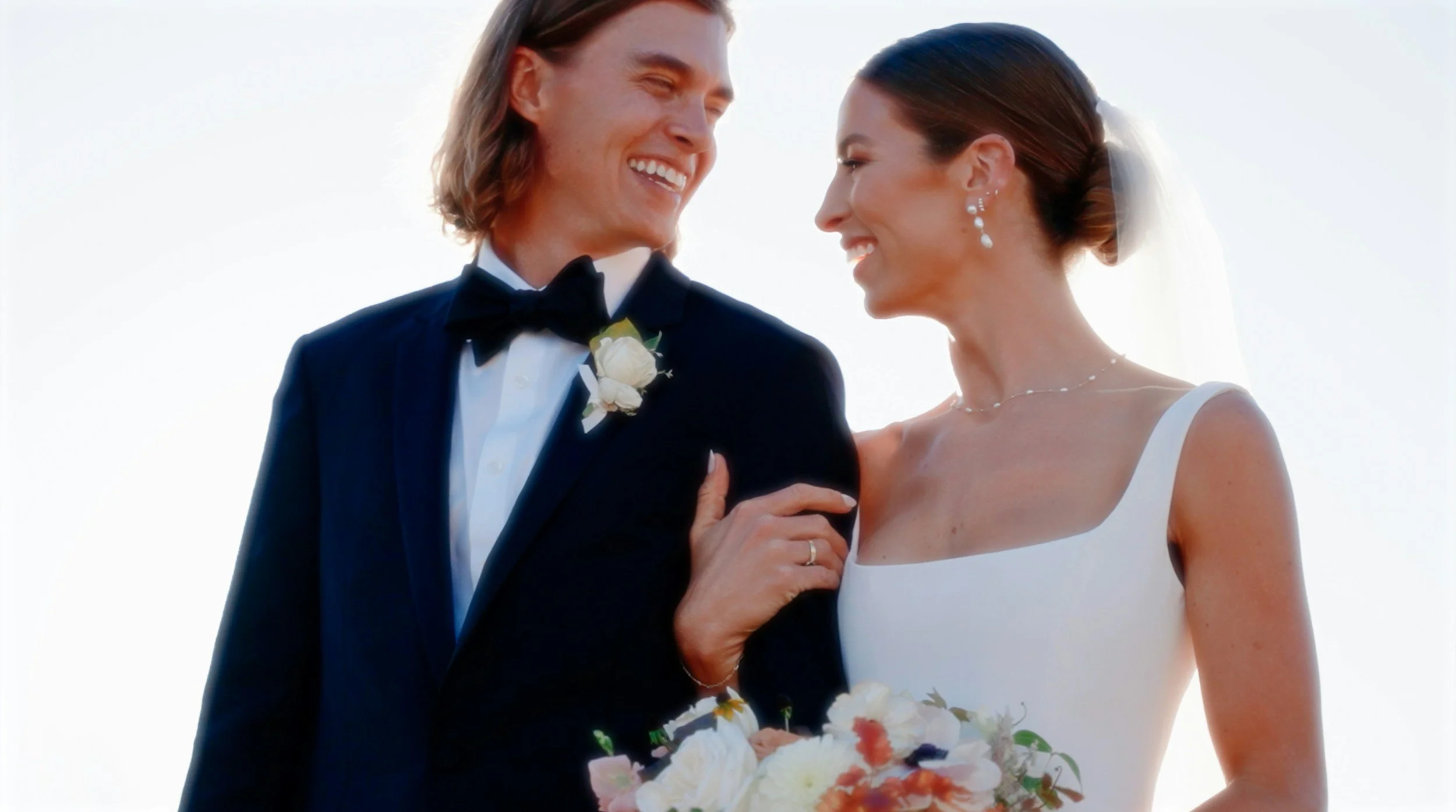 A newlywed couple smiling at each other outdoors during sunset, with the groom in a black tuxedo and the bride in a white wedding dress, holding a bouquet of flowers.