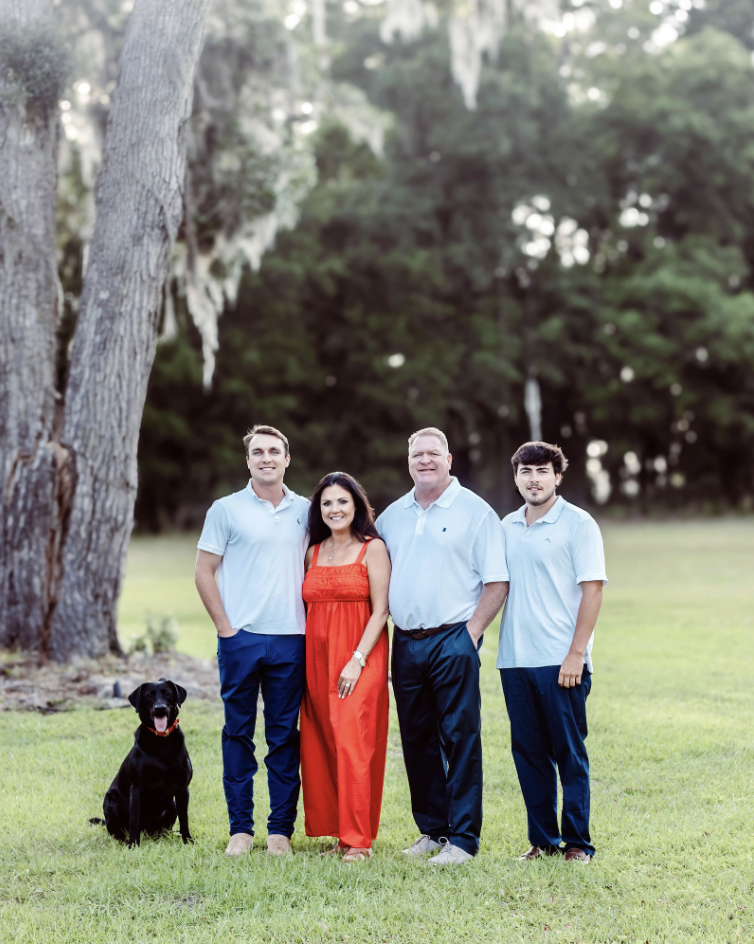 The Jones Family of four with a black dog standing on grass in a park with large trees in the background, posing for a photo. The mother is in a red dress, the father and two sons are in white shirts and blue pants.