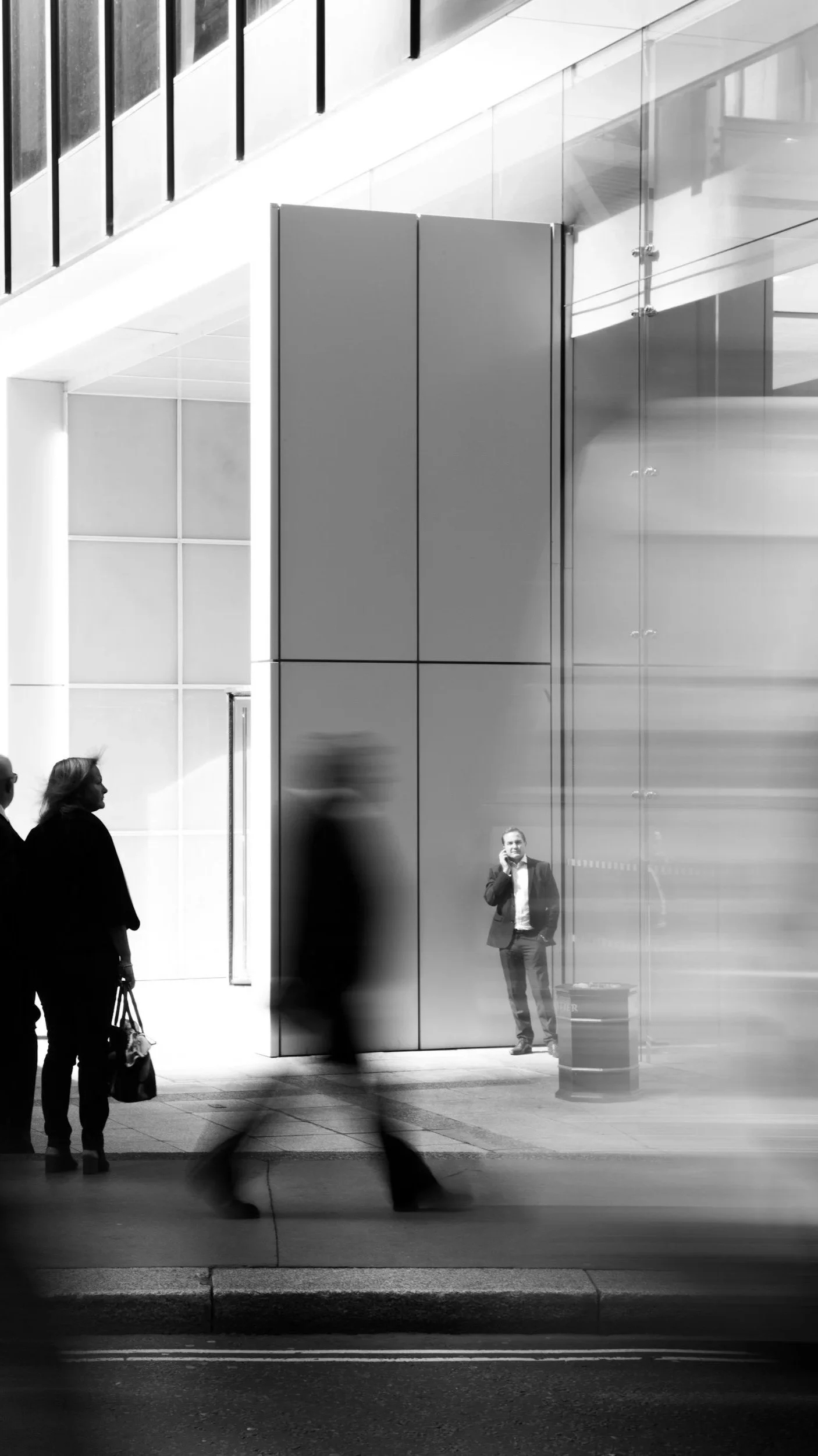 People walking past a modern building entrance with glass and metallic panels, a man in a suit talking on his phone, and others blurred in motion.