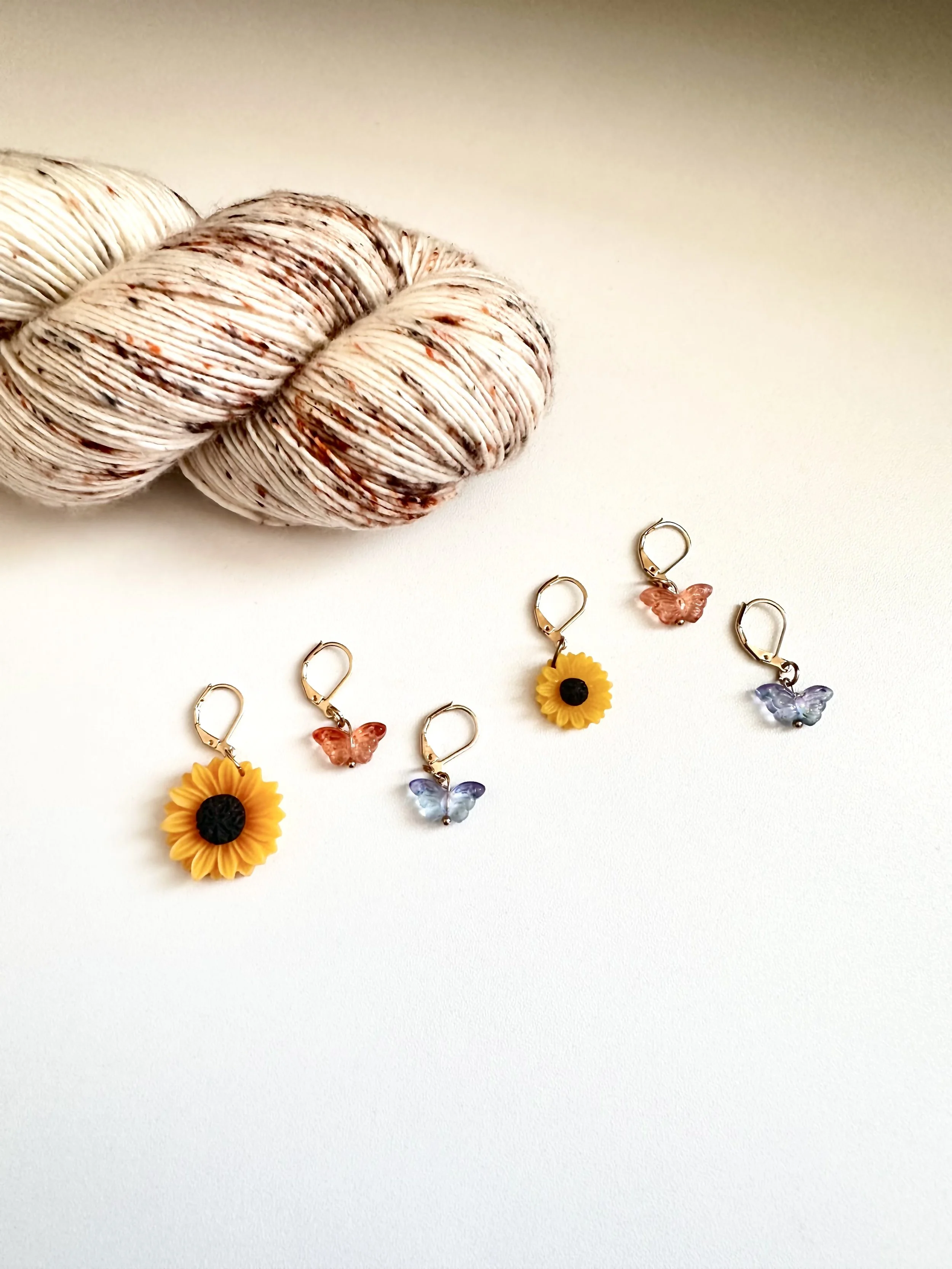 Colorful handmade knitting and crochet stitch markers with sunflower and butterfly charms, arranged on a white surface, with a skein of speckled yarn in the background.