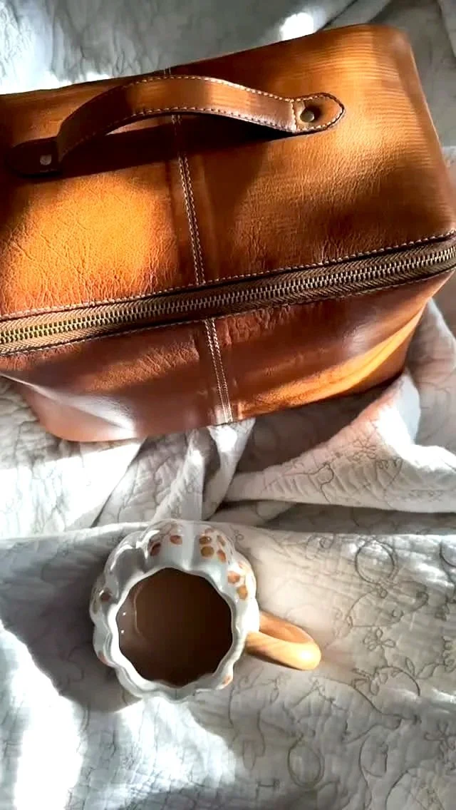 A brown leather travel bag with a zipper on a quilted fabric surface, and a ceramic mug filled with a hot beverage, with a floral design, a handle, and pouring spout.