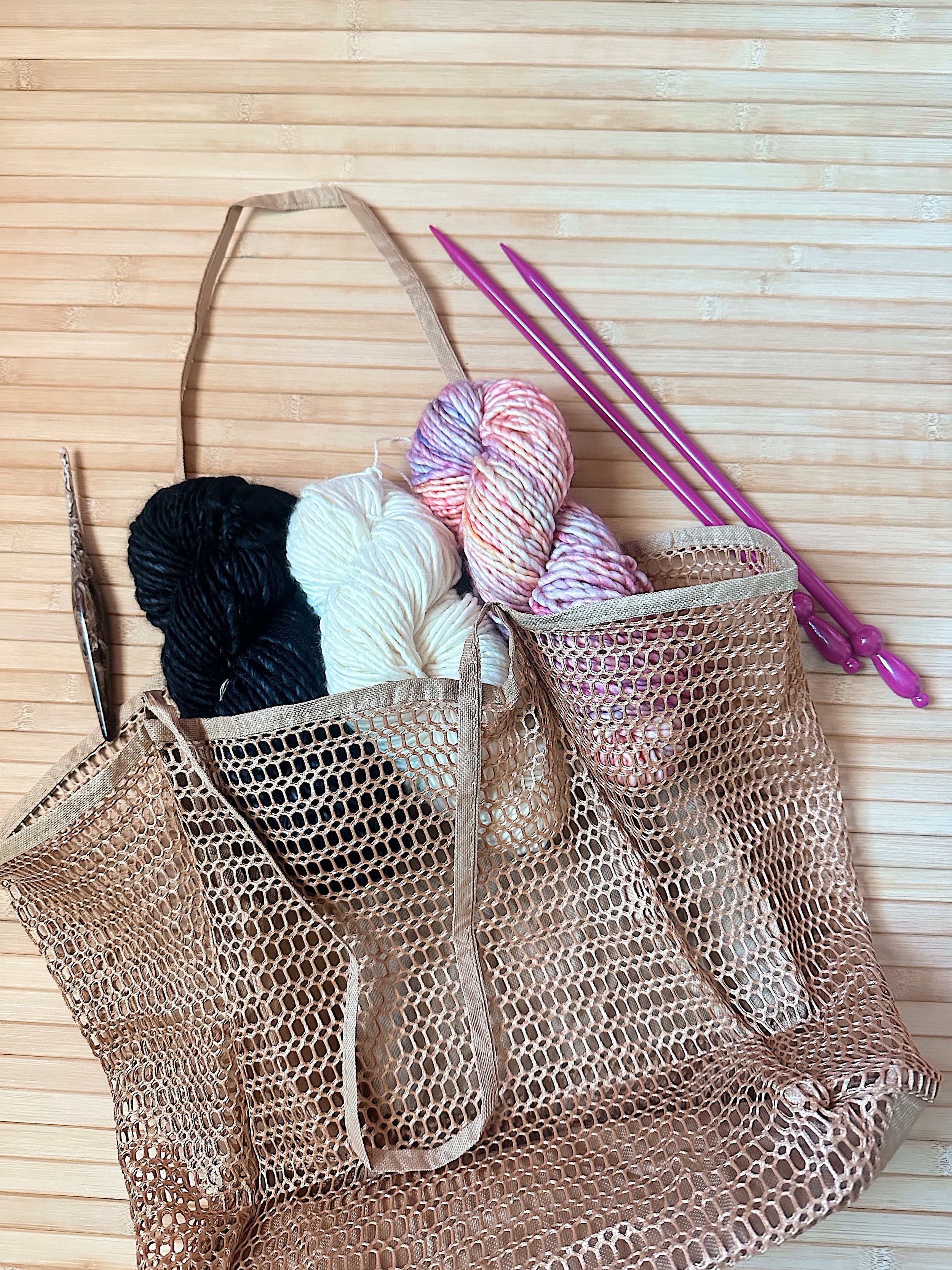 A mesh shopping tote bag containing several balls of yarn in various colors, including black, white, and pink, with two purple knitting needles resting on top, placed on a wooden surface.