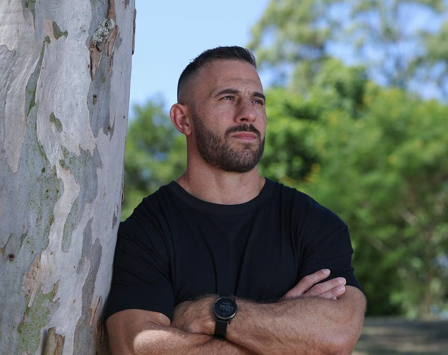Dave Kramer, wearing a black shirt and a watch, stands outdoors next to a tree with peeling bark, with green trees and a blue sky in the background.