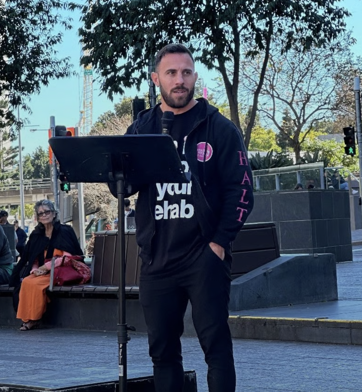 Dave Kramer speaking into a microphone outdoors at a What Were You Wearing rally, standing behind a music stand, wearing a black sweatshirt with various texts and logos, with trees and a traffic light in the background.