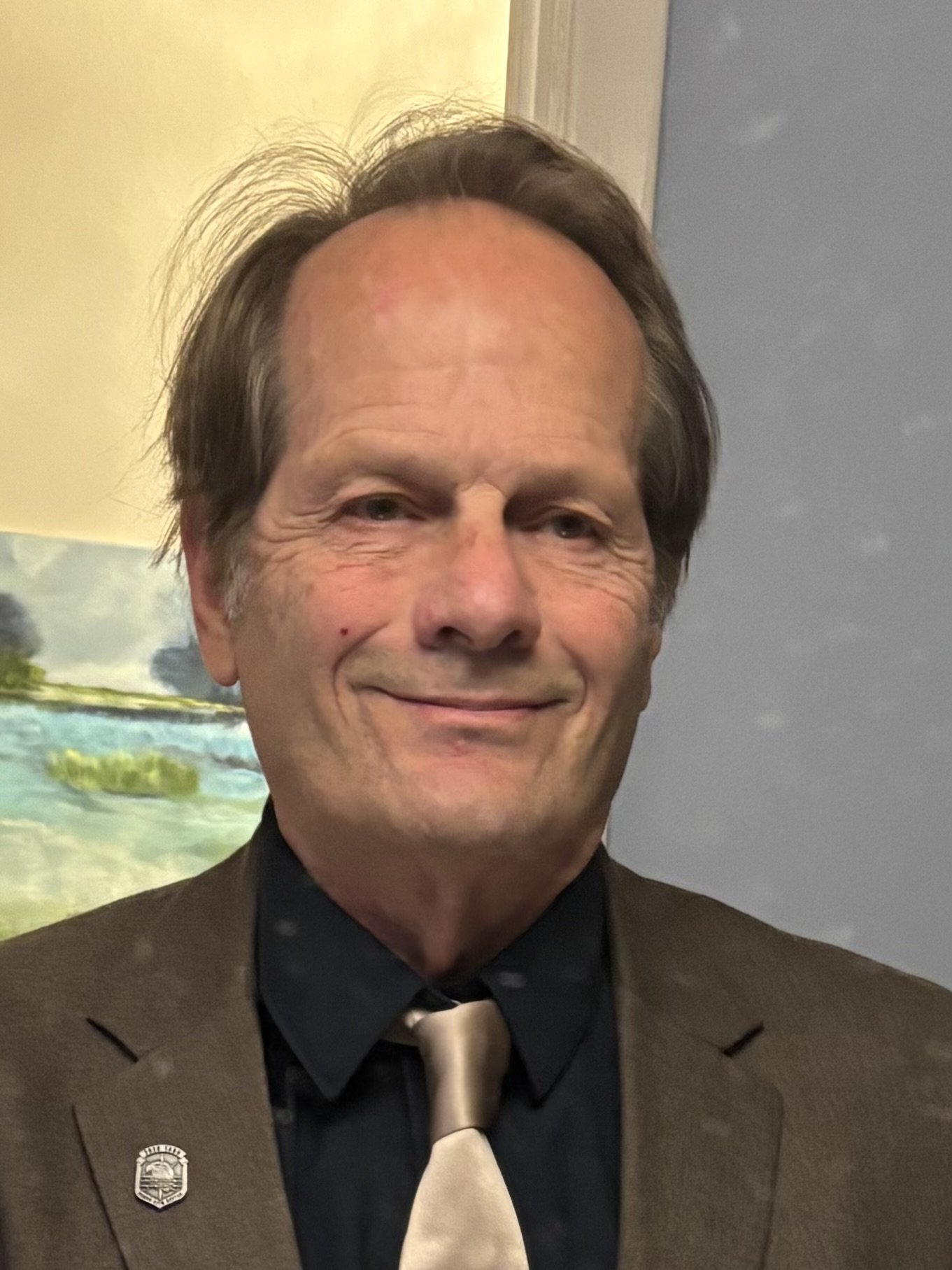 A smiling man with brown hair and wrinkles, wearing a brown blazer and a black shirt with a beige tie, standing indoors in front of a wall with a painting of a river and trees.