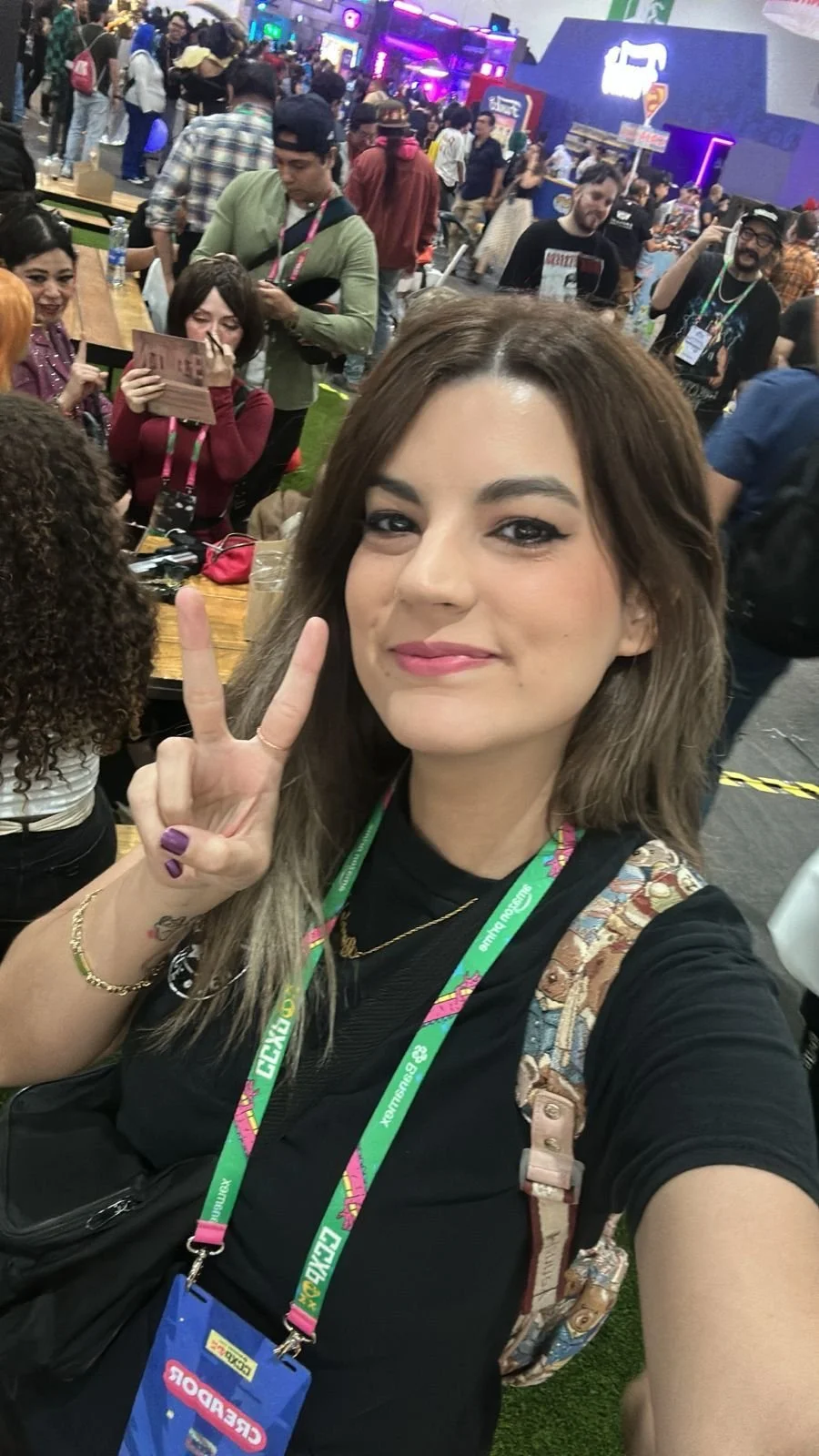 Joven mujer en un evento o convención, haciendo un símbolo de paz con la mano, rodeada de muchas personas y luces de colores.