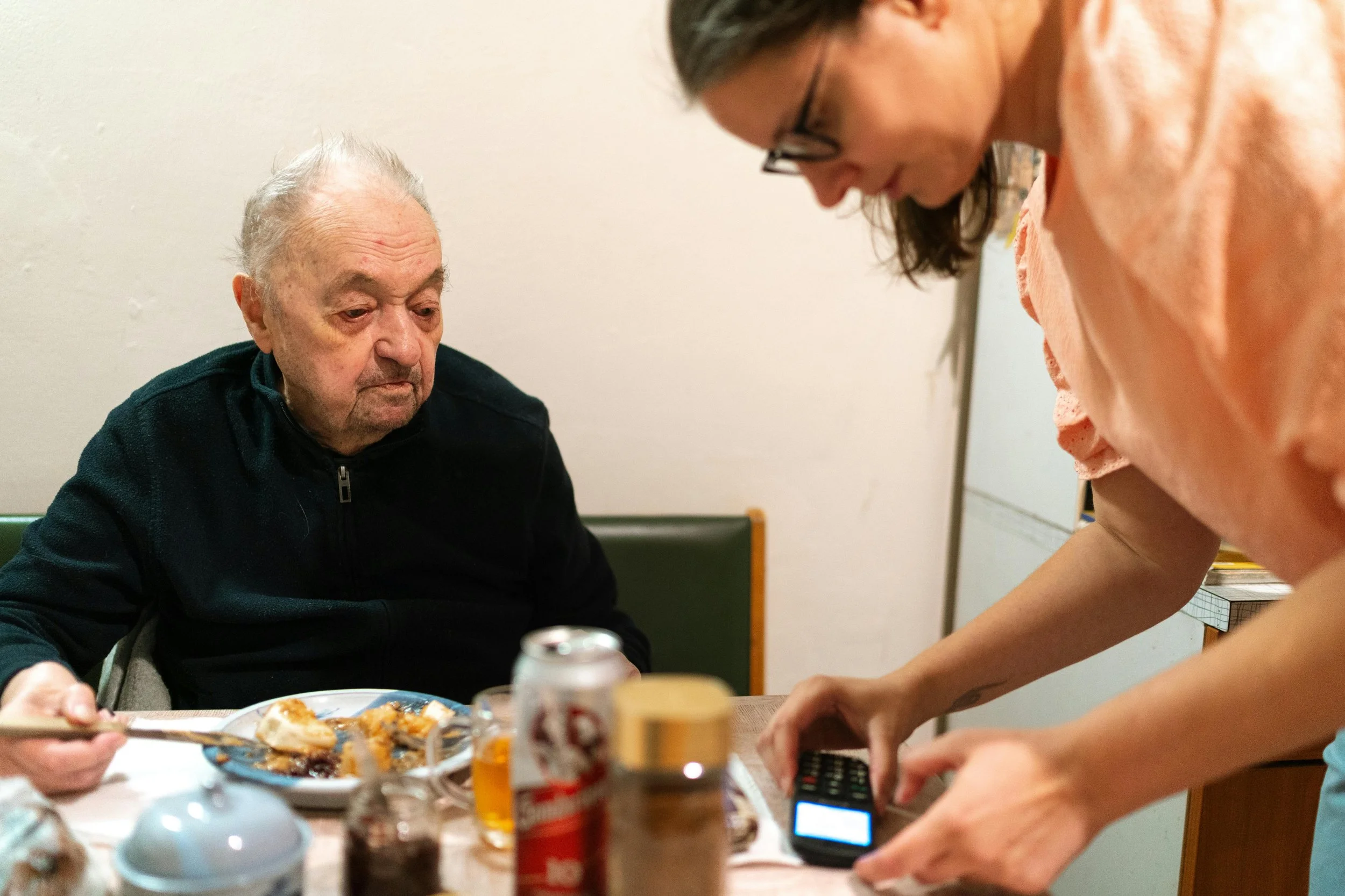An elderly man with gray hair and a black zip-up sweater sitting at a table, looking down, with a woman in glasses and a pink shirt leaning over the table, assisting him. The table has various food items, drinks, and condiments on it.