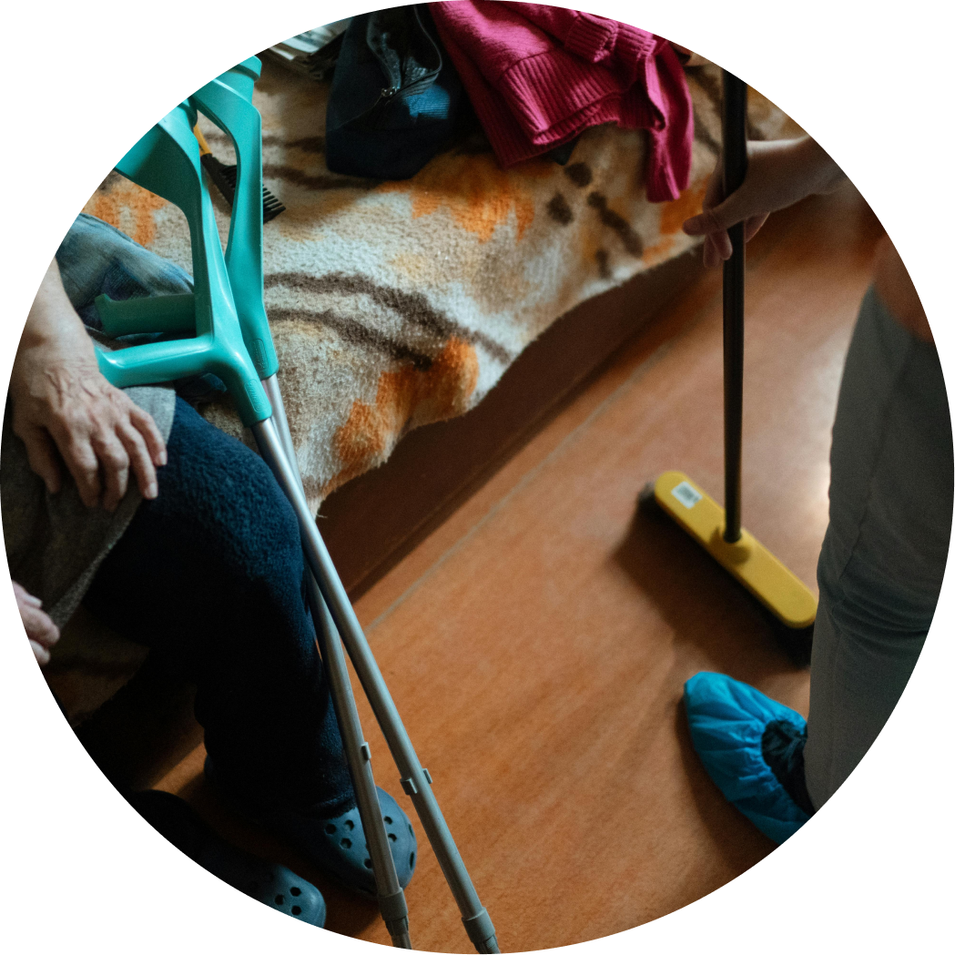 A person standing on a wooden floor wearing blue shoe covers, next to an elderly person sitting on a couch with a walker nearby. The person standing is holding a yellow stick with a black handle, possibly a broom or mop. On the couch, there are some clothes, including a pink sleeve and a blue item. The elderly person has a hand resting on their lap.