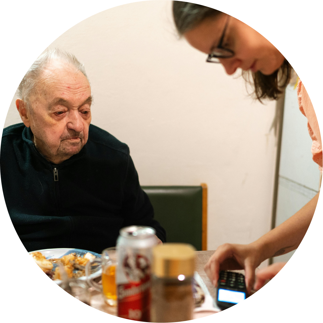 An elderly man with gray hair and a black jacket sitting at a table, looking down at another person who is standing and using a calculator or remote control, with food, a can, and medication on the table.