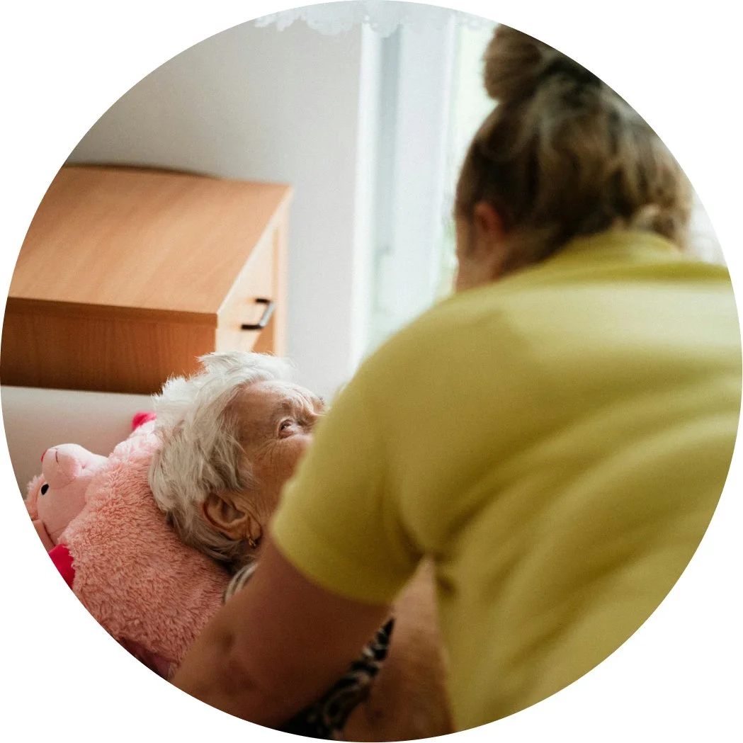 An elderly woman lying in bed with a pink stuffed animal, looking at a caregiver who is leaning over her and talking to her.