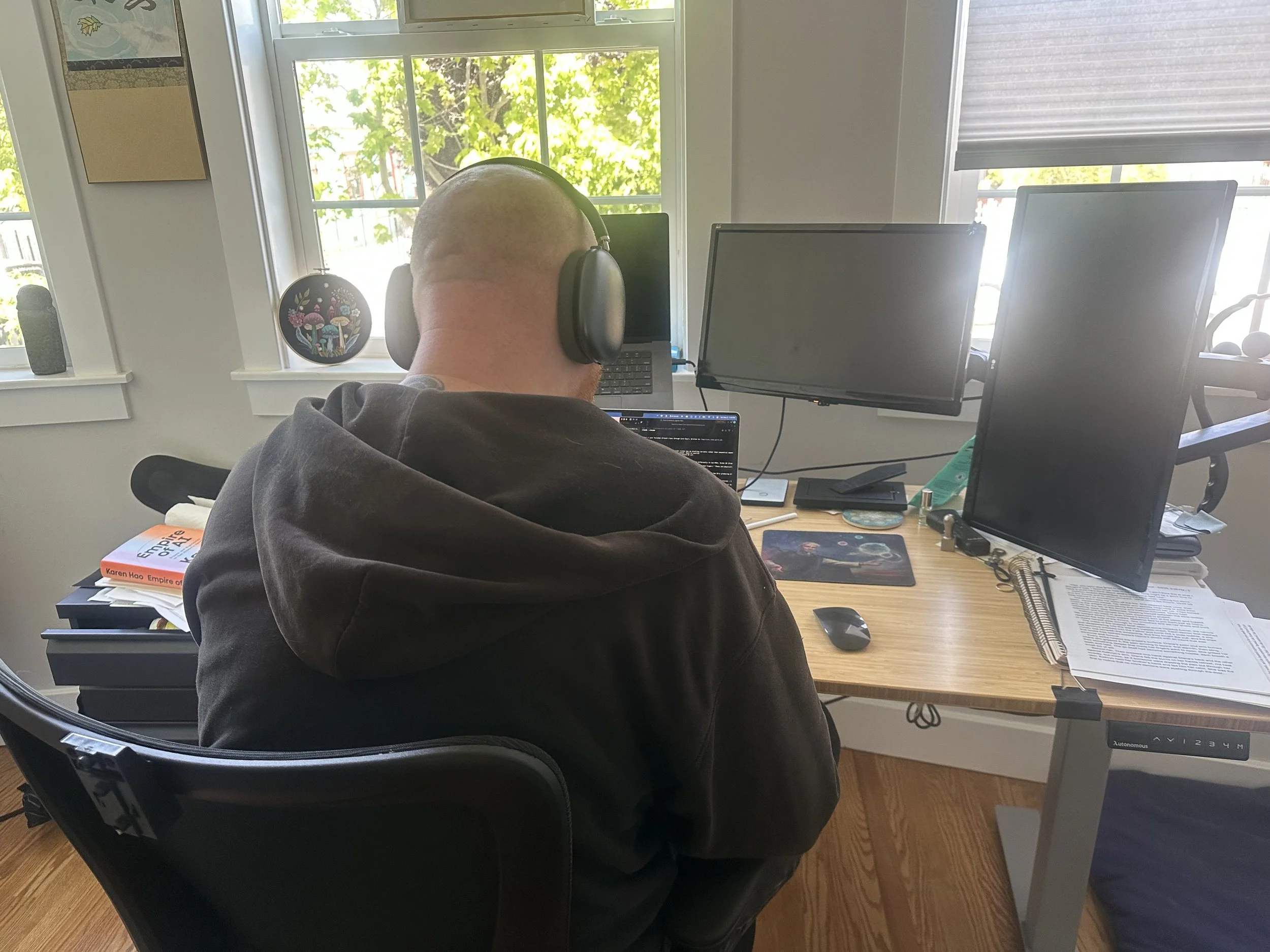 A man sitting at a cluttered desk using a laptop, with two monitors, wearing large headphones. A notebook and various papers are on the desk, with a window showing green trees outside behind him.
