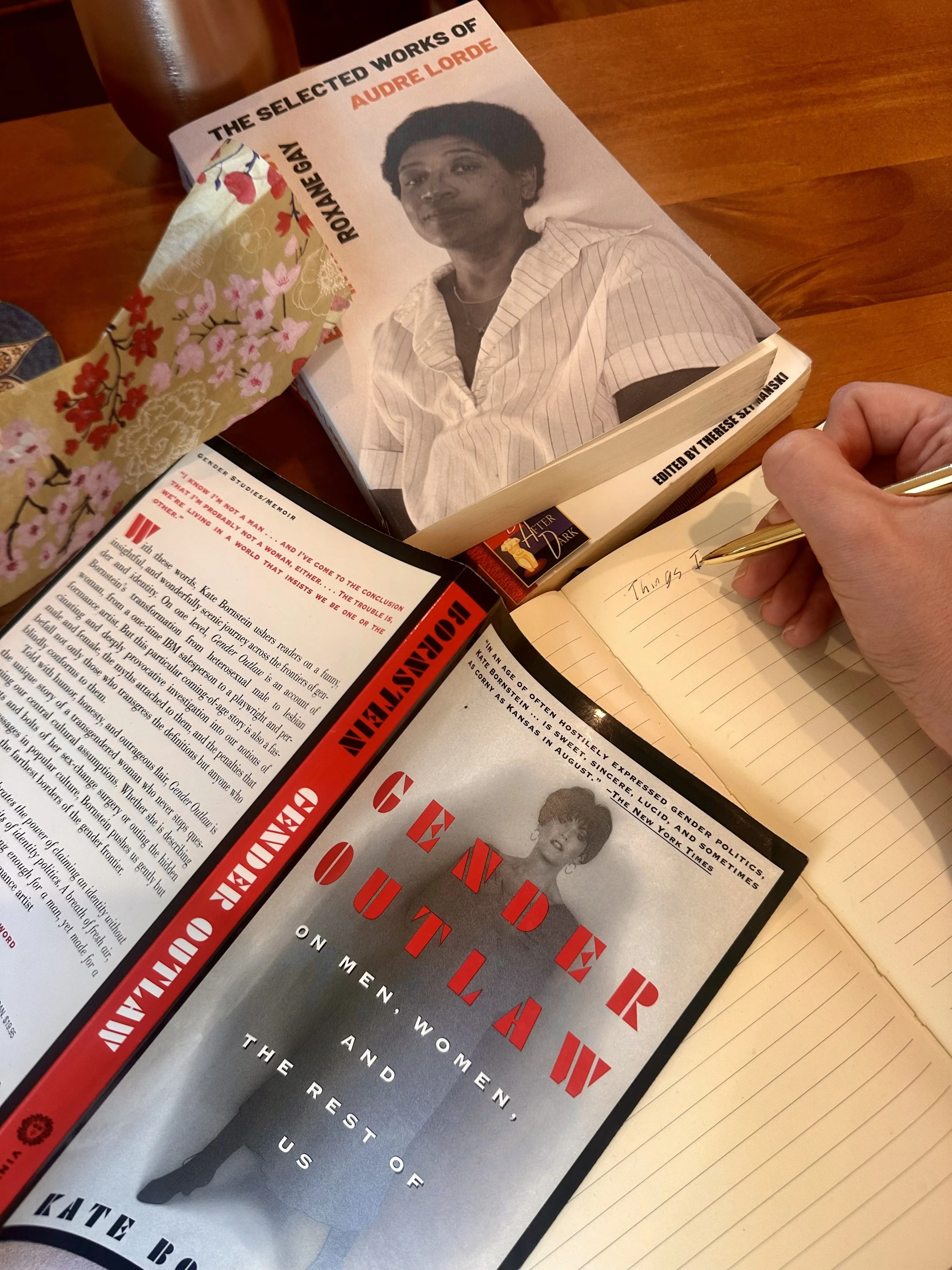 A person is writing on a lined notebook with a gold pen. On the table, there is a magazine titled "The Gendered Brain" and a book about Audrey Lorde, along with a black and white photo of Audrey Lorde on the cover. There is a magazine page with a black and white photograph of a woman with short hair and a striped shirt, and text about her.