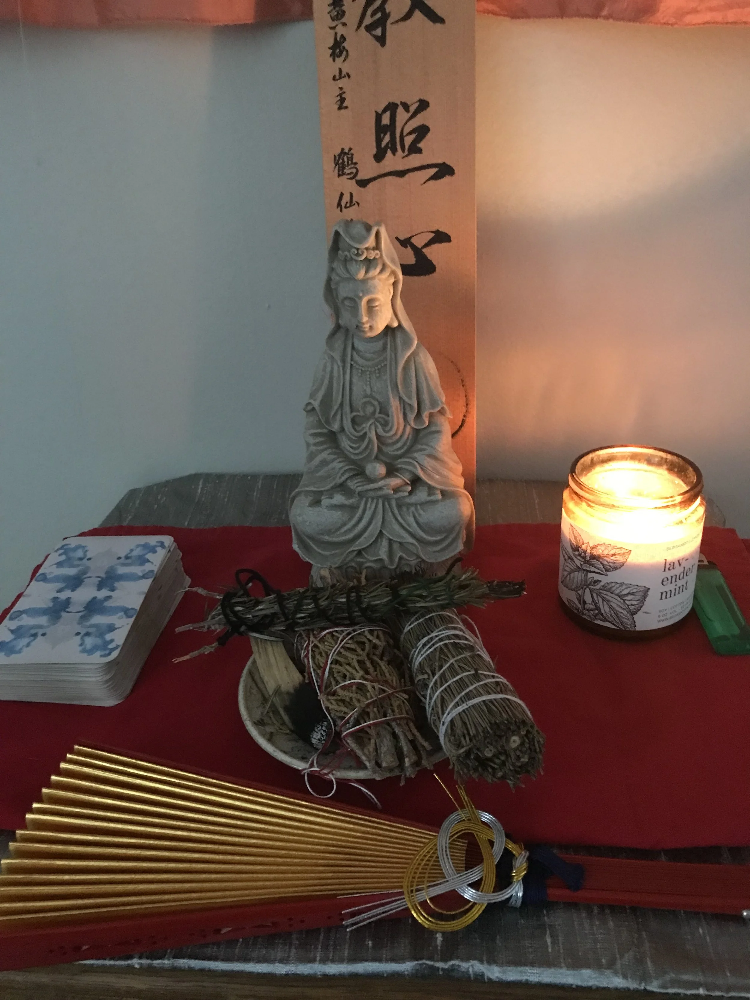 A small statue of a deity or spiritual figure with ornate attire, seated with legs crossed, surrounded by candles, bundles of incense sticks, and golden sticks, on a wooden surface at a spiritual or religious altar.