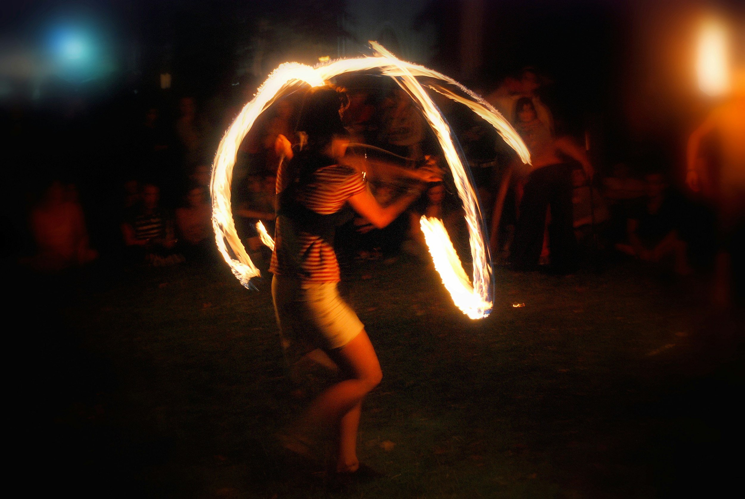 A woman performing a fire dance at night, creating a circle of fire with a flaming object, with onlookers in the background.