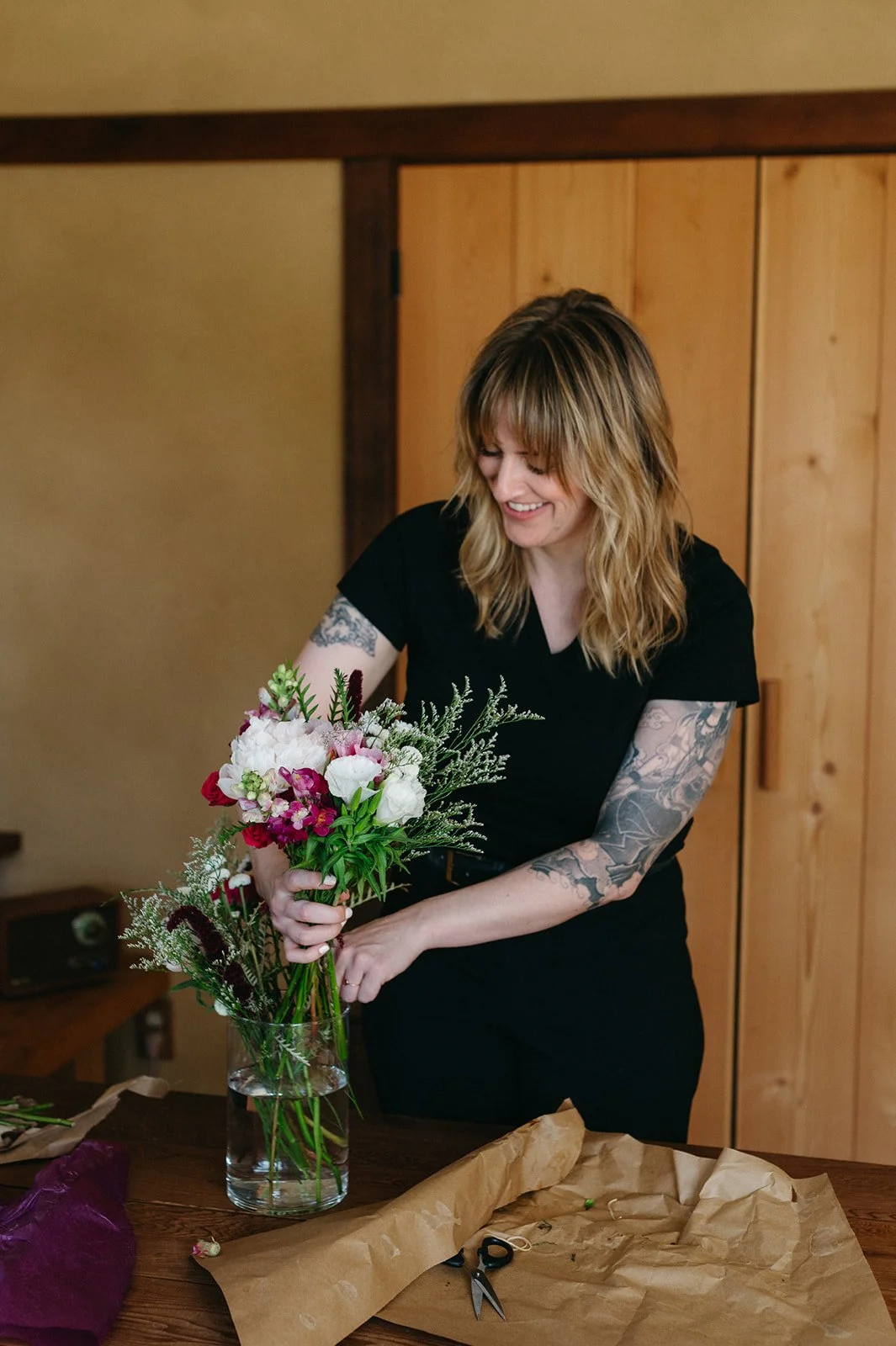 A woman with shoulder-length blonde hair and tattoos on her arms arranging flowers in a glass vase. She is smiling and wearing a black shirt, standing at a wooden table with flower arranging supplies.