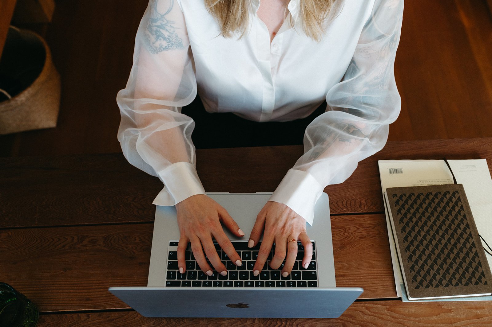 A woman wearing a white see-through blouse working on a silver MacBook at a wooden desk, with notebooks and papers on the right side.