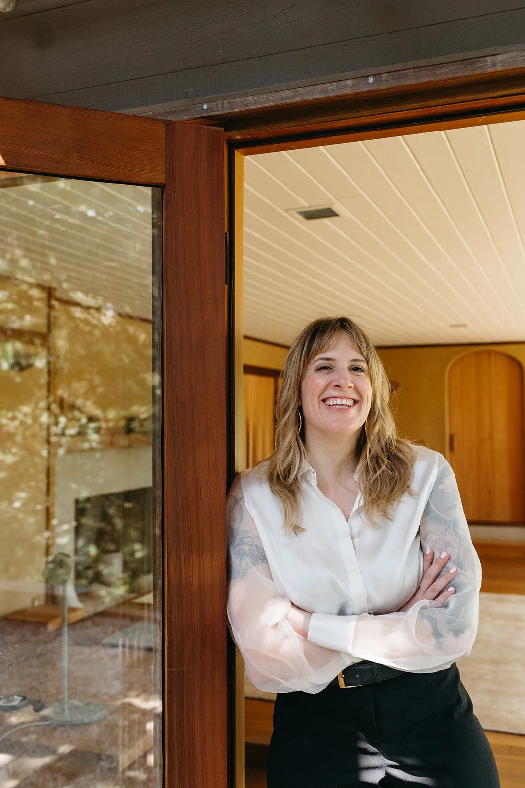 A woman smiling with arms crossed, standing partially outside a wooden house, with an indoor panel ceiling and a warm-toned interior.