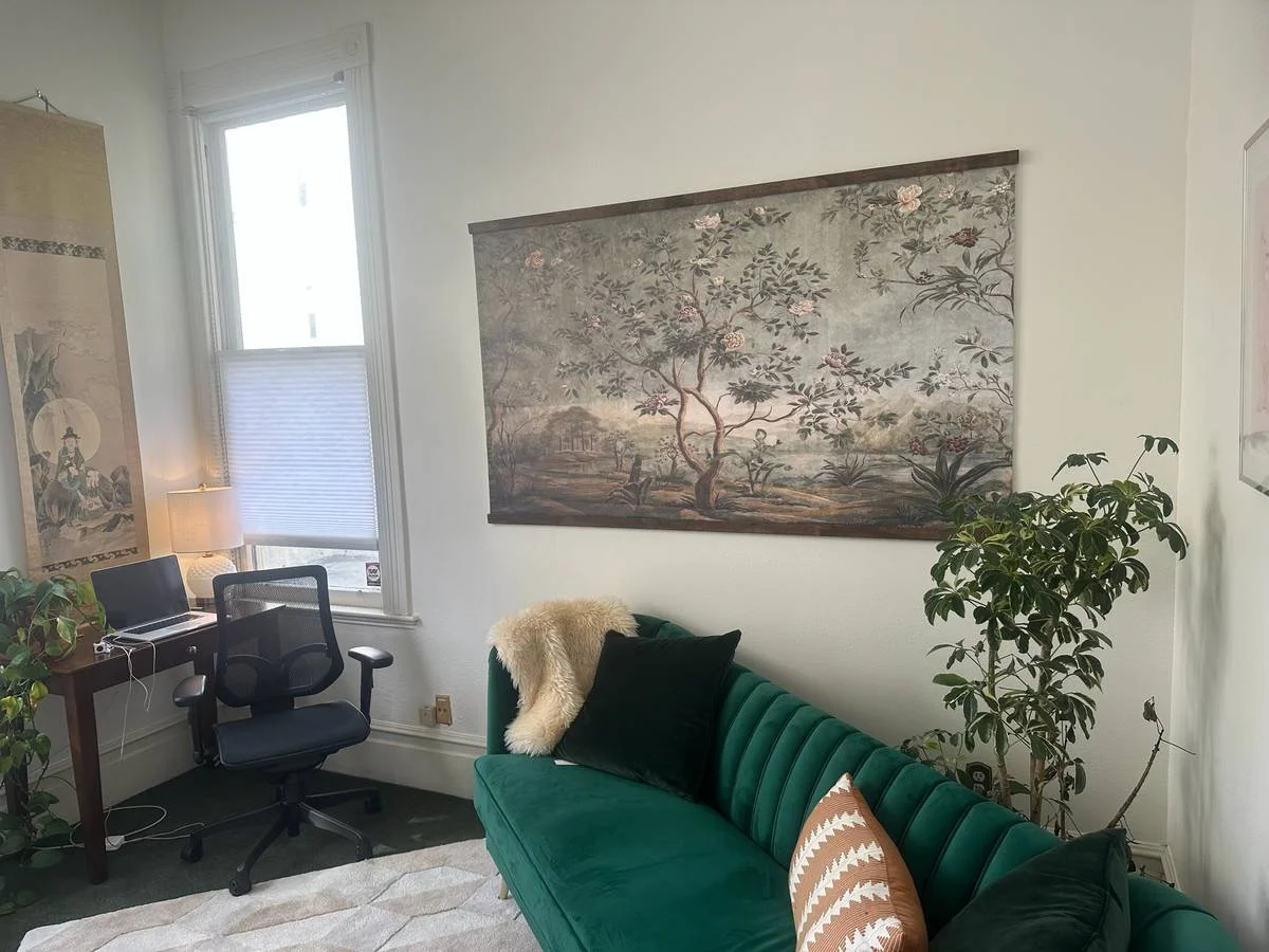 a cozy living room corner with a green velvet sofa decorated with cushions, a large tapestry of a tree on the wall, a desk with a laptop, a swivel chair, and a window with blinds, plants, and a lamp.