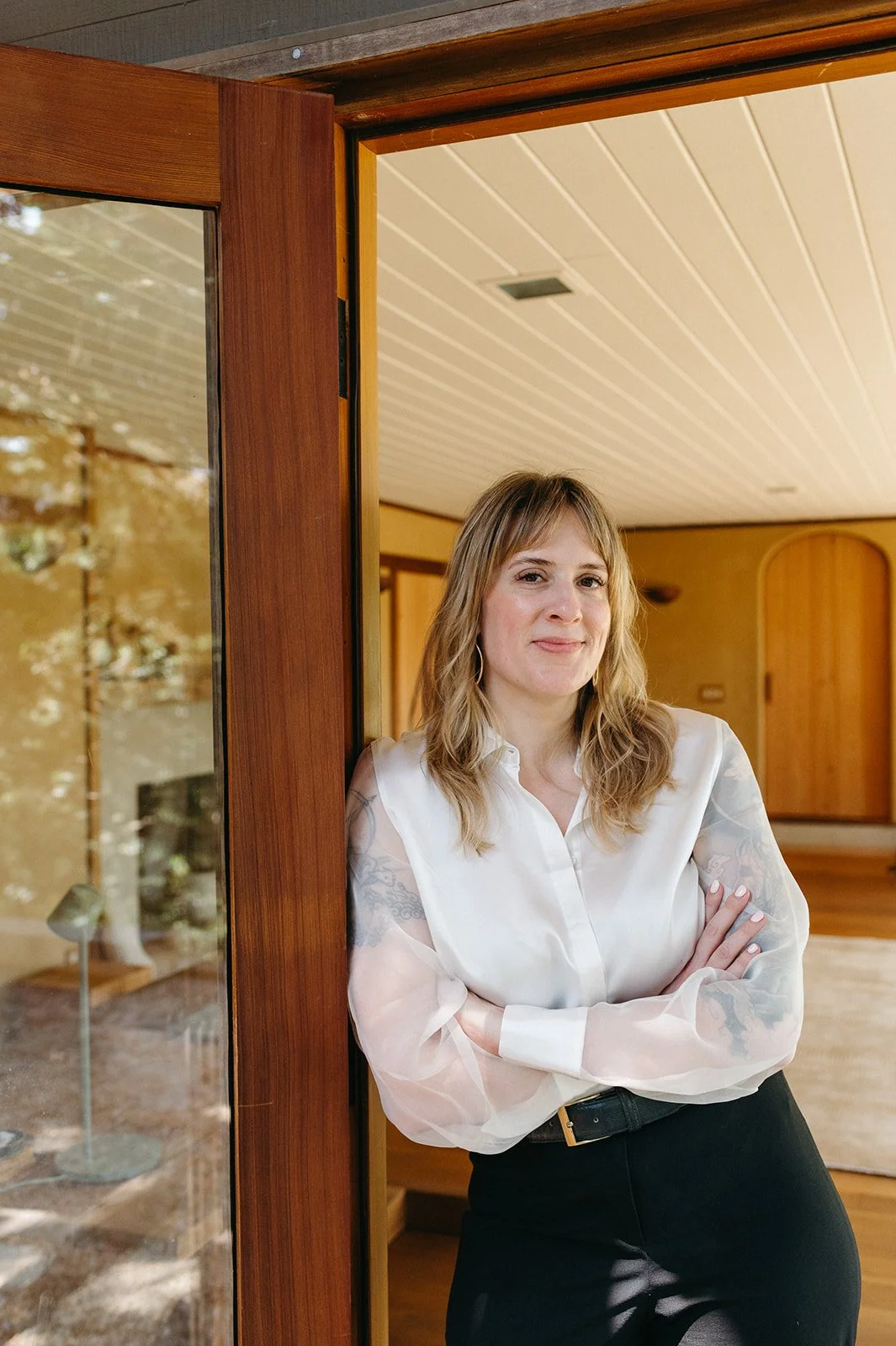 Woman standing with arms crossed inside a room, leaning against a wooden doorway, wearing a white blouse and black pants, with a wooden interior and glass door behind her.