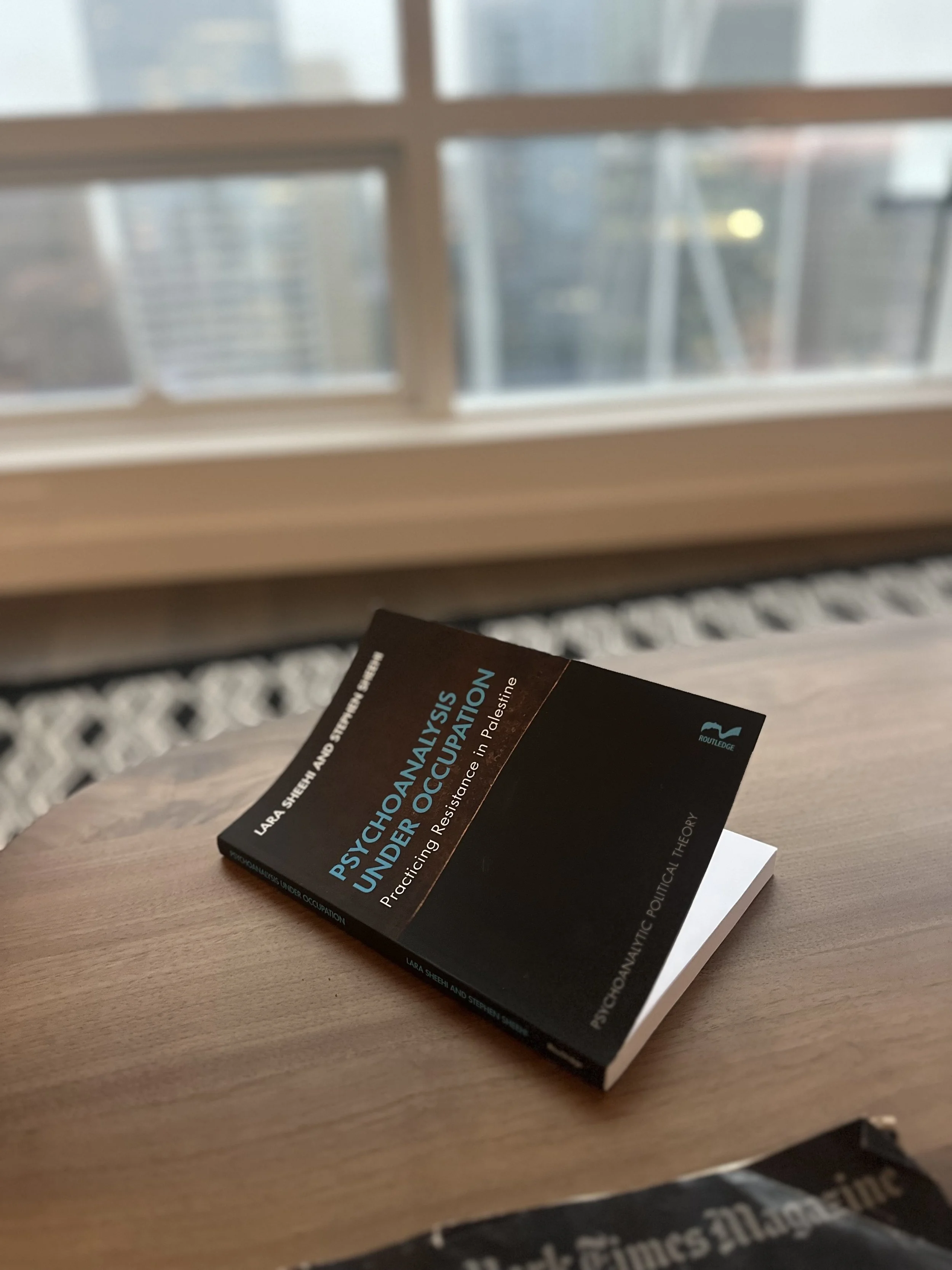 Close-up of a book titled "Psychopathology Under Occupation" resting on a wooden table near a window with blurry cityscape in the background.