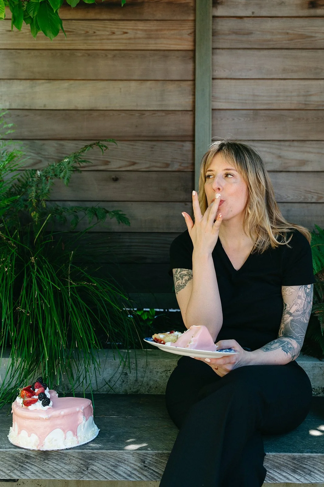 A young woman with blonde hair and tattoos on her arms is sitting outdoors against a wooden fence, eating a slice of cake and holding a plate with cake in her lap. There is a pink cake decorated with berries on the ground in front of her.
